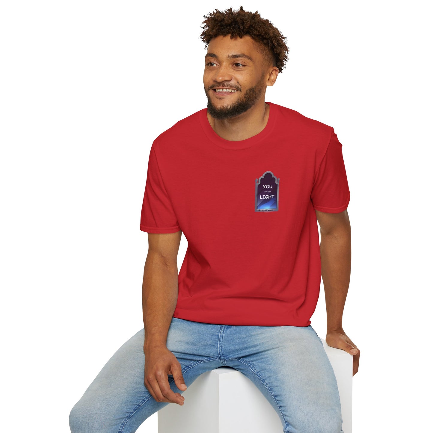 A man in a red shirt, sitting on a cube, showcasing the YOU ARE THE LIGHT small Mirror Unisex T-shirt. Classic fit, 100% cotton tee with smooth surface for premium printing.