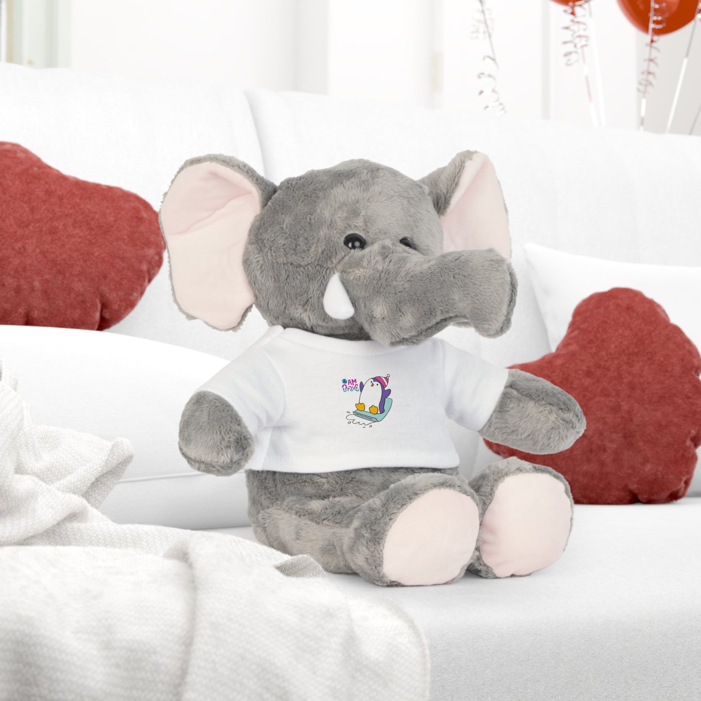Cool Stuff Stuffies Elephant with 'I am Brave' Penguin design on a removable white t-shirt, sitting on a couch. Part of a comforting plush toy collection.