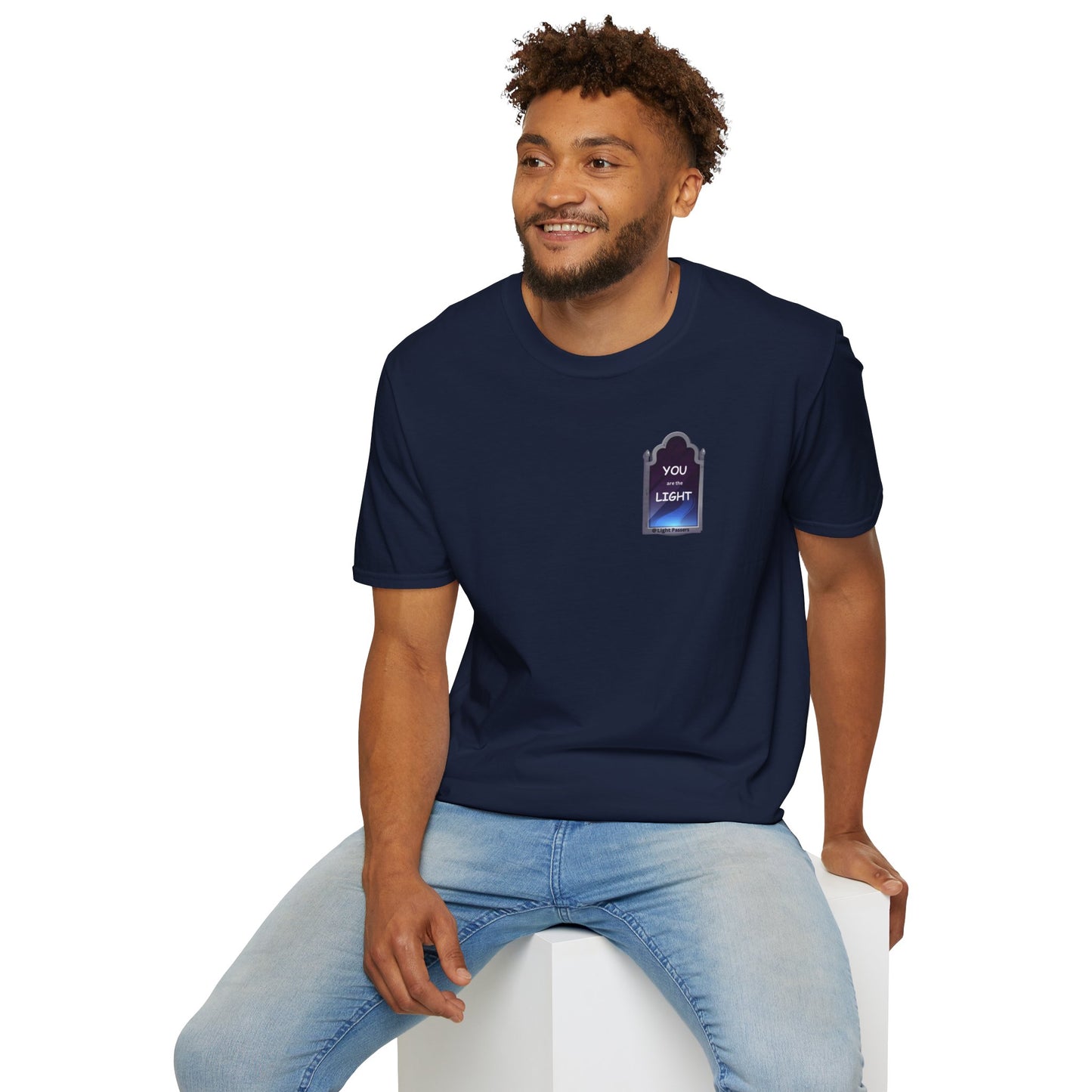 A man in a blue shirt sits on a white cube, showcasing the YOU ARE THE LIGHT small Mirror Unisex T-shirt. The tee offers a classic fit, 100% cotton, tear-away label, and durable shoulder tape for comfort.