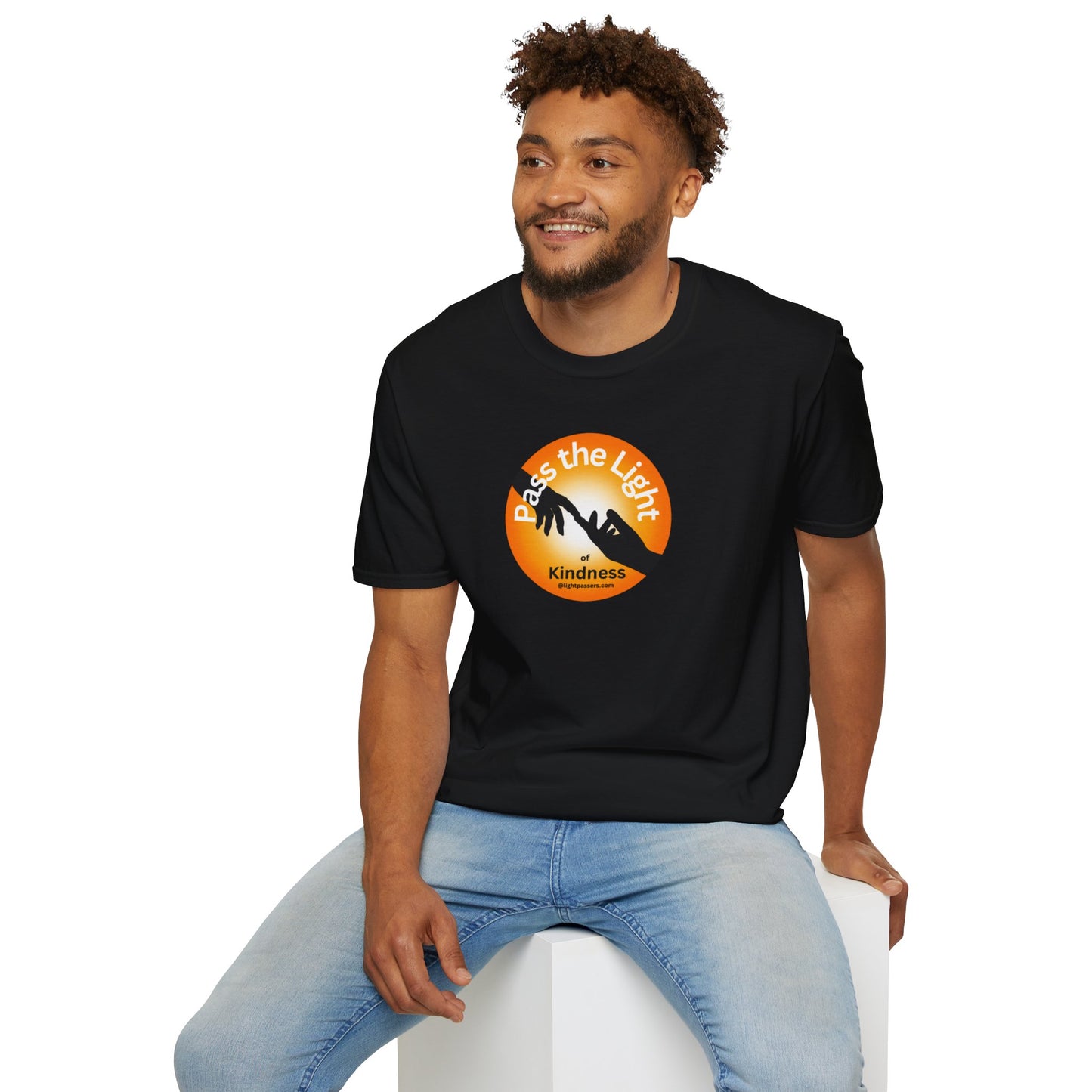 A man in a black shirt sits on a cube, showcasing the Golden Ball Pass the Light Unisex T-shirt. Close-ups reveal logo details and the man's smile and curly hair.