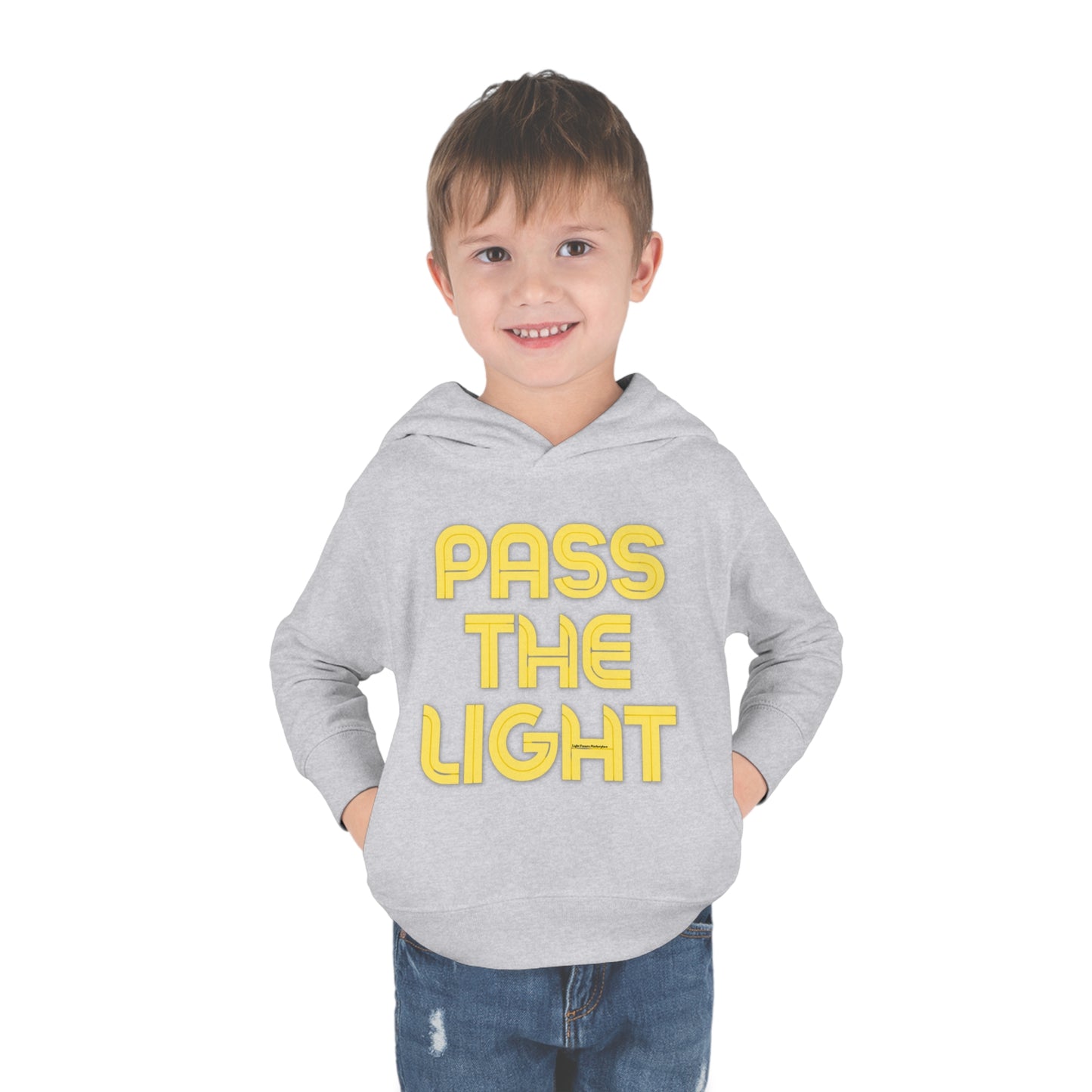 A smiling boy in jeans, wearing a Pass the Light Toddler Hooded Sweatshirt with hands in pockets. Features jersey-lined hood, cover-stitched details, side-seam pockets for cozy comfort.