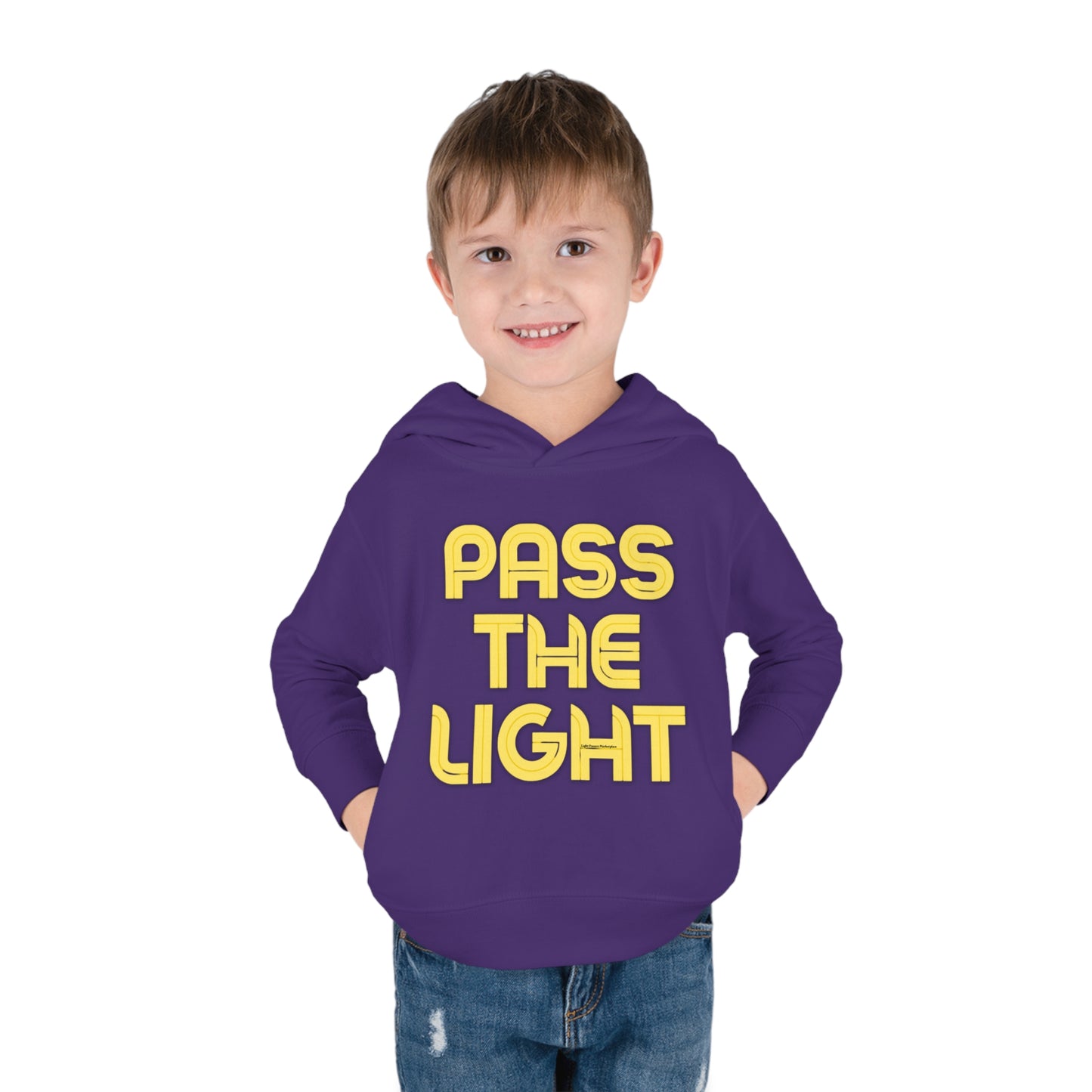 A boy in a Pass the Light Toddler Hooded Sweatshirt with yellow text, hands in pockets, close-ups of clothing details like pants and face, showcasing durable design with jersey-lined hood, cover-stitched accents, and side-seam pockets for cozy wear.