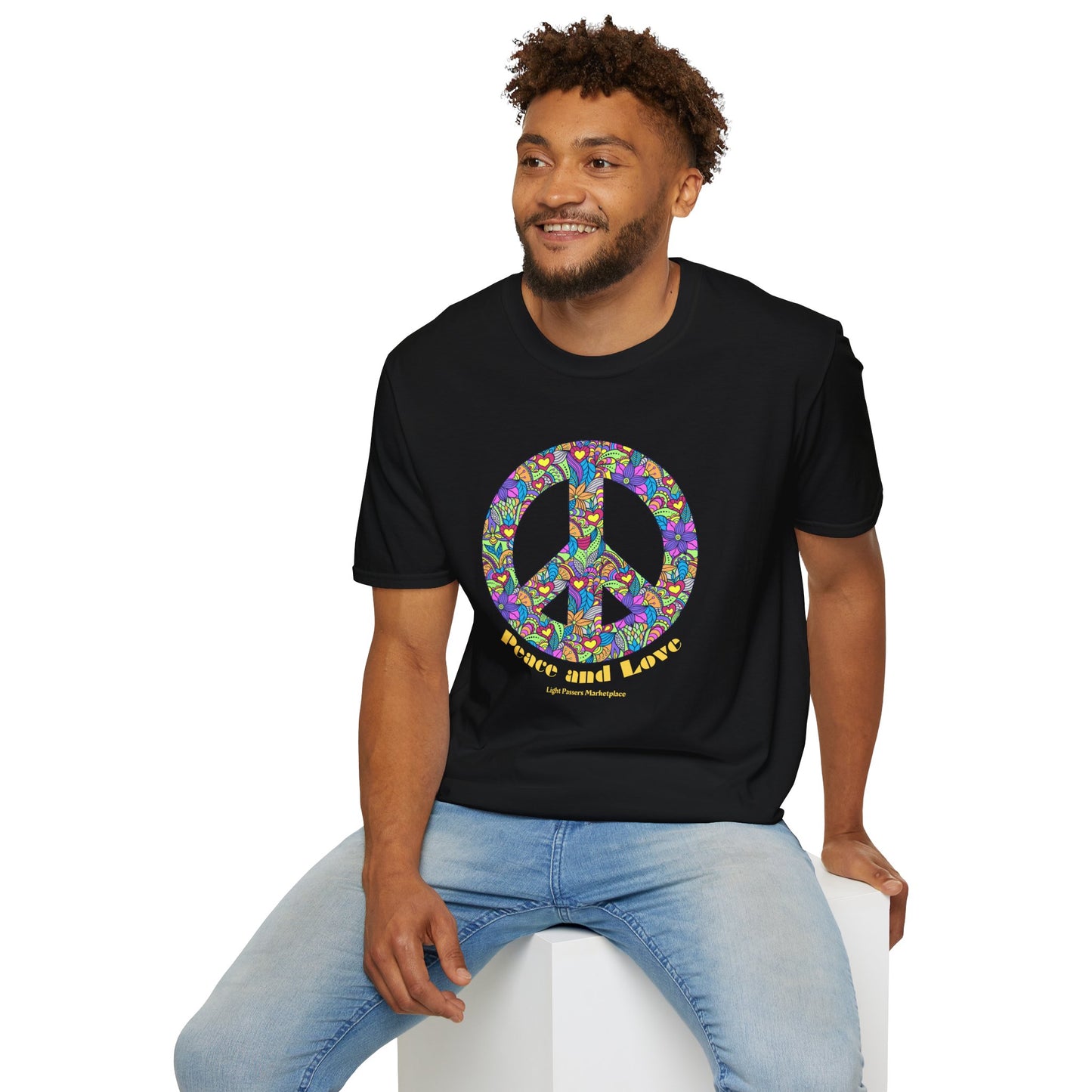 A man sitting on a cube, wearing a black shirt with a peace sign, showcasing the Peace sign with flowers Unisex T-shirt. Made of soft 100% cotton, featuring twill tape shoulders and a ribbed collar.