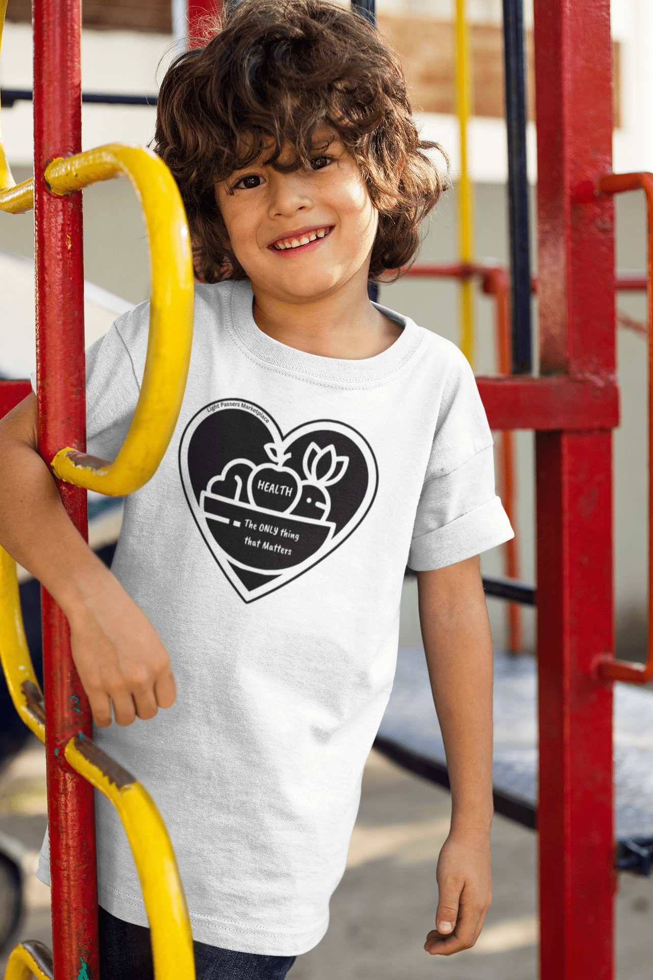 A child smiles on a playground, wearing the Fruit Basket Youth T-shirt. Made with 100% cotton, featuring twill tape shoulders and ribbed collar for durability and comfort.