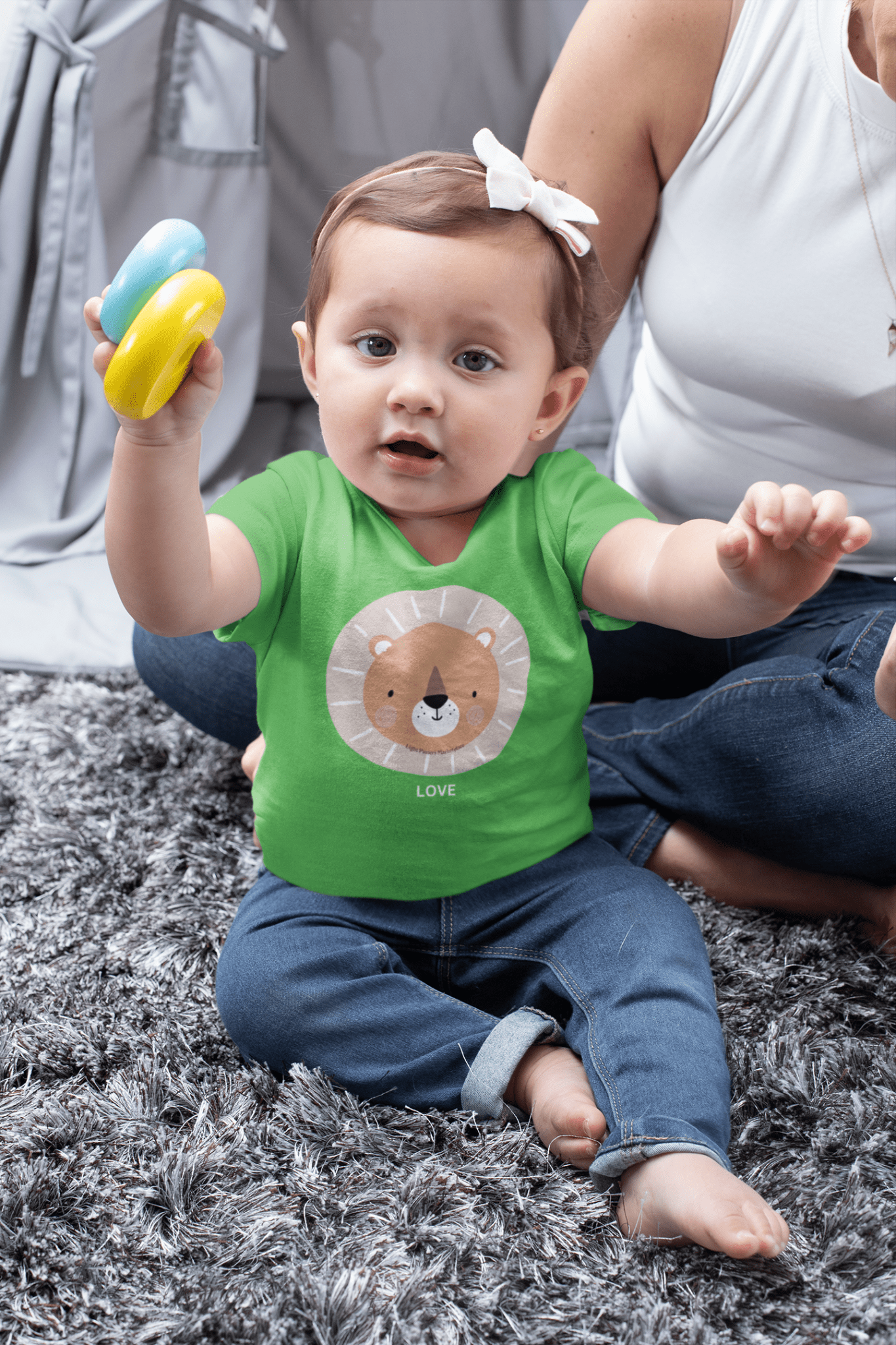 A baby wearing a green shirt sits on the floor, holding a lion face toy. This infant fine jersey tee offers comfort and durability with side seams, ribbed knitting, and taped shoulders.