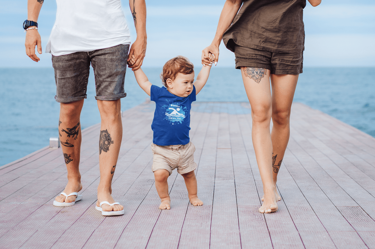 A baby in a blue shirt walks on a wooden surface, holding hands with a man and woman. Swim for Health Baby T-shirts offer comfort and durability with side seams, ribbed knitting, and taped shoulders.