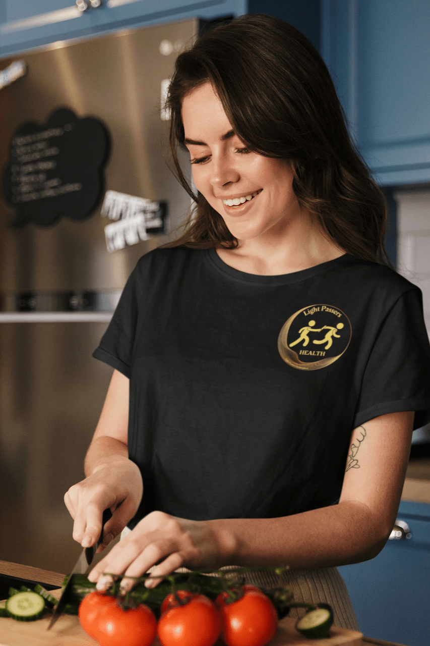 A woman in a kitchen cutting vegetables, wearing a black shirt, embodies the comfort of the Light Passers Relay Health small Logo Unisex T-Shirt. Made of soft 100% cotton, featuring twill tape shoulders and a ribbed collar.