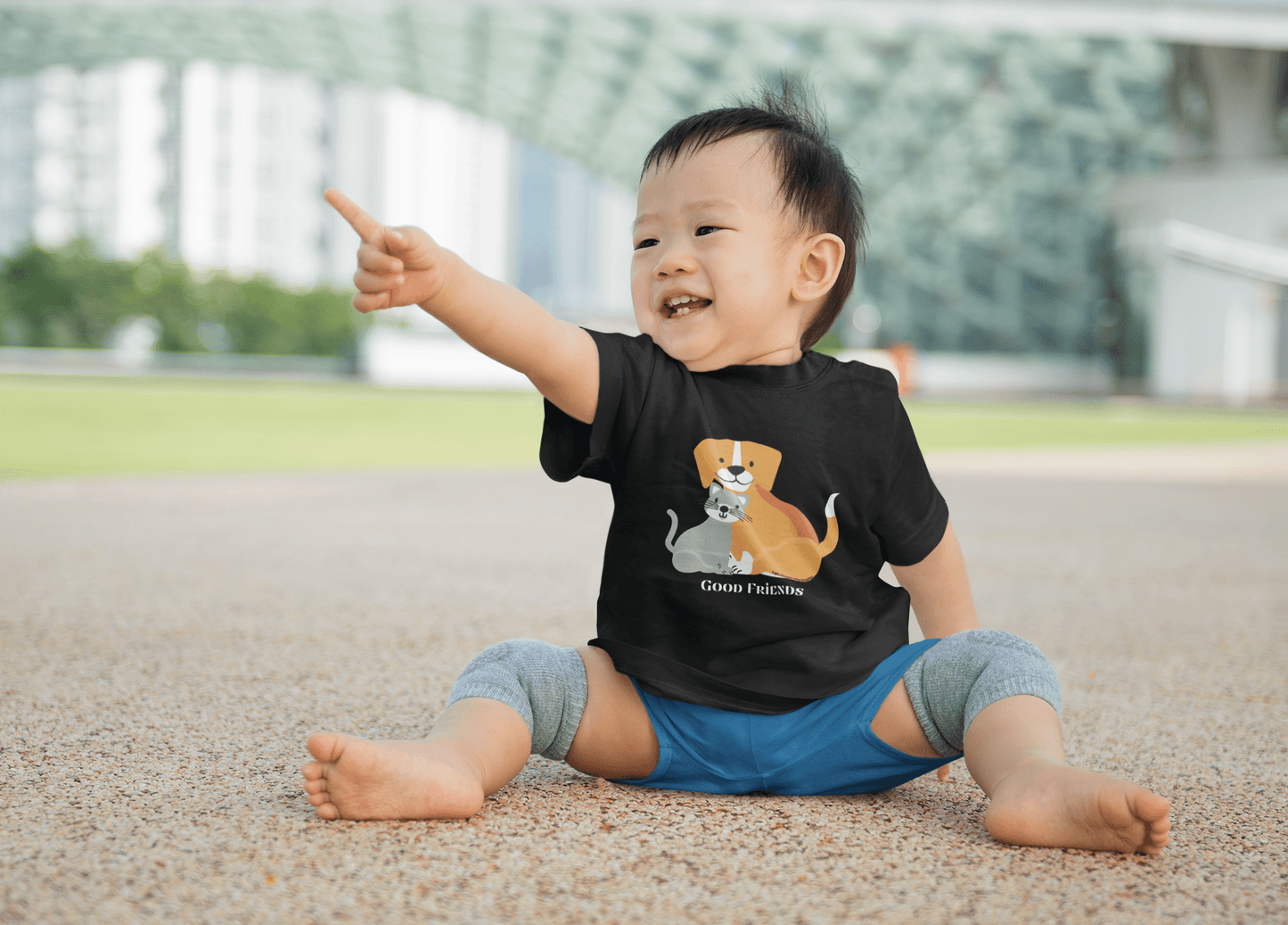 A toddler in a black tee with a cat and dog design, pointing, showcasing comfort and durability with side seams, ribbed knitting, and taped shoulders.