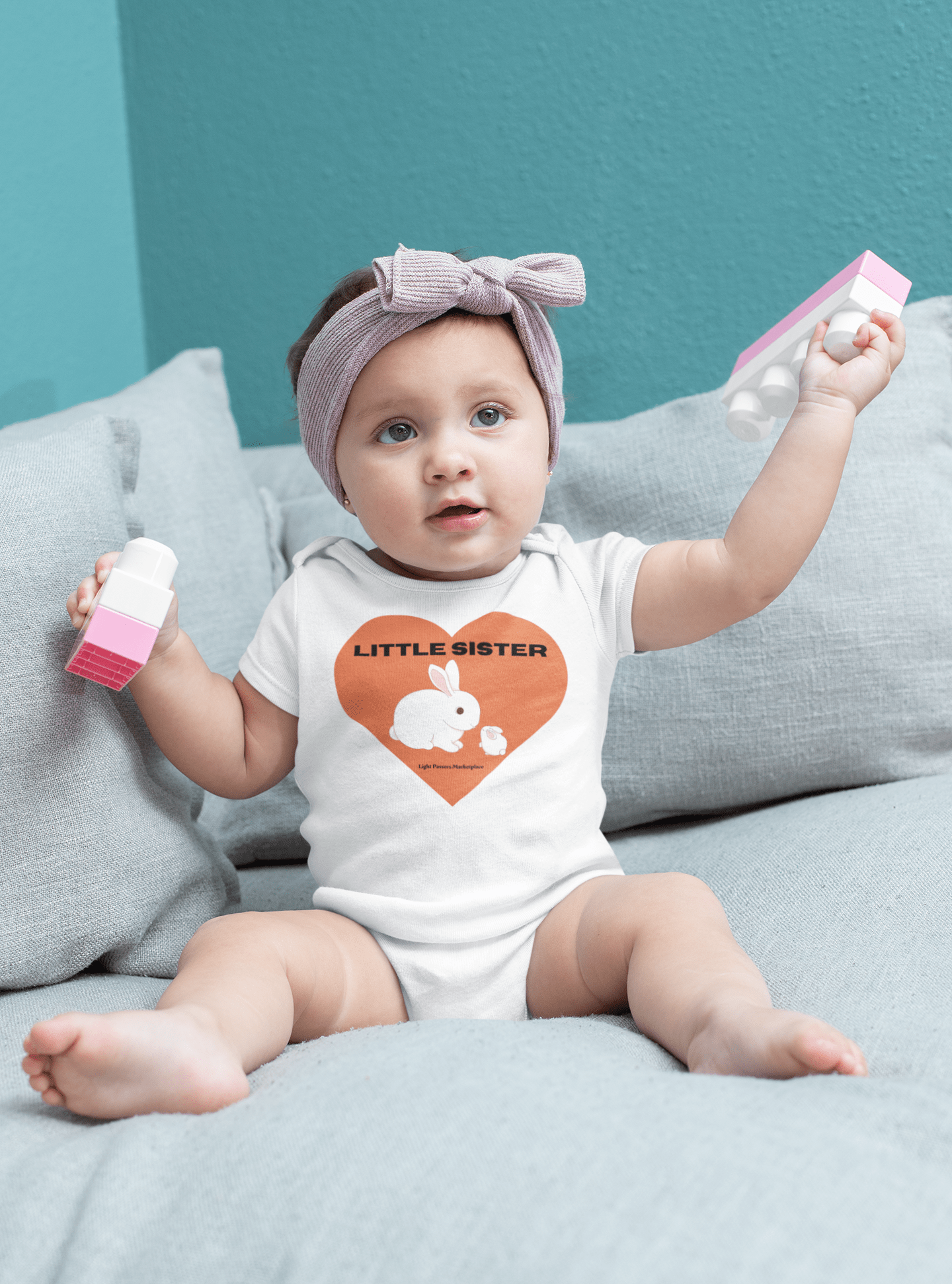 A baby in a Little Sister Onesie, holding a toy. 100% organic cotton, OEKO-TEX® certified. Bottom snap closure, ribbed legs for secure fit. Printed care label, Onesies® Brand tag.