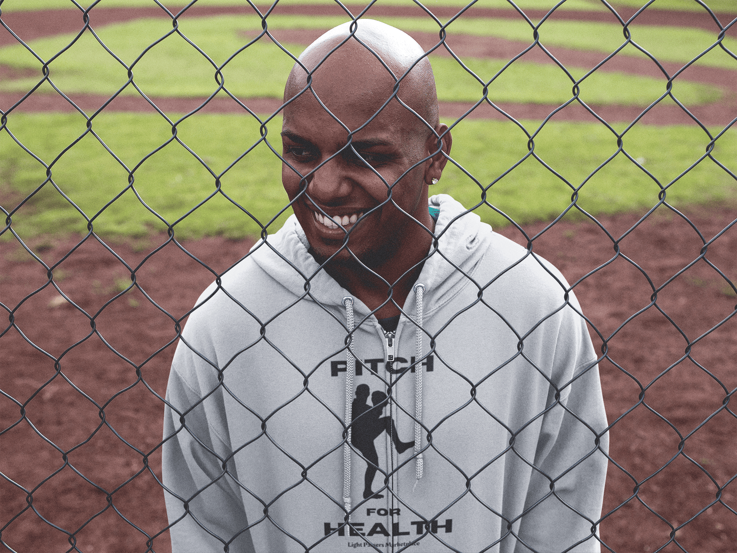 A man smiles behind a chain-link fence, embodying comfort in the Pitch For Health Unisex Hooded Sweatshirt. Thick cotton-polyester blend, kangaroo pocket, and color-matched drawstring offer warmth and style.