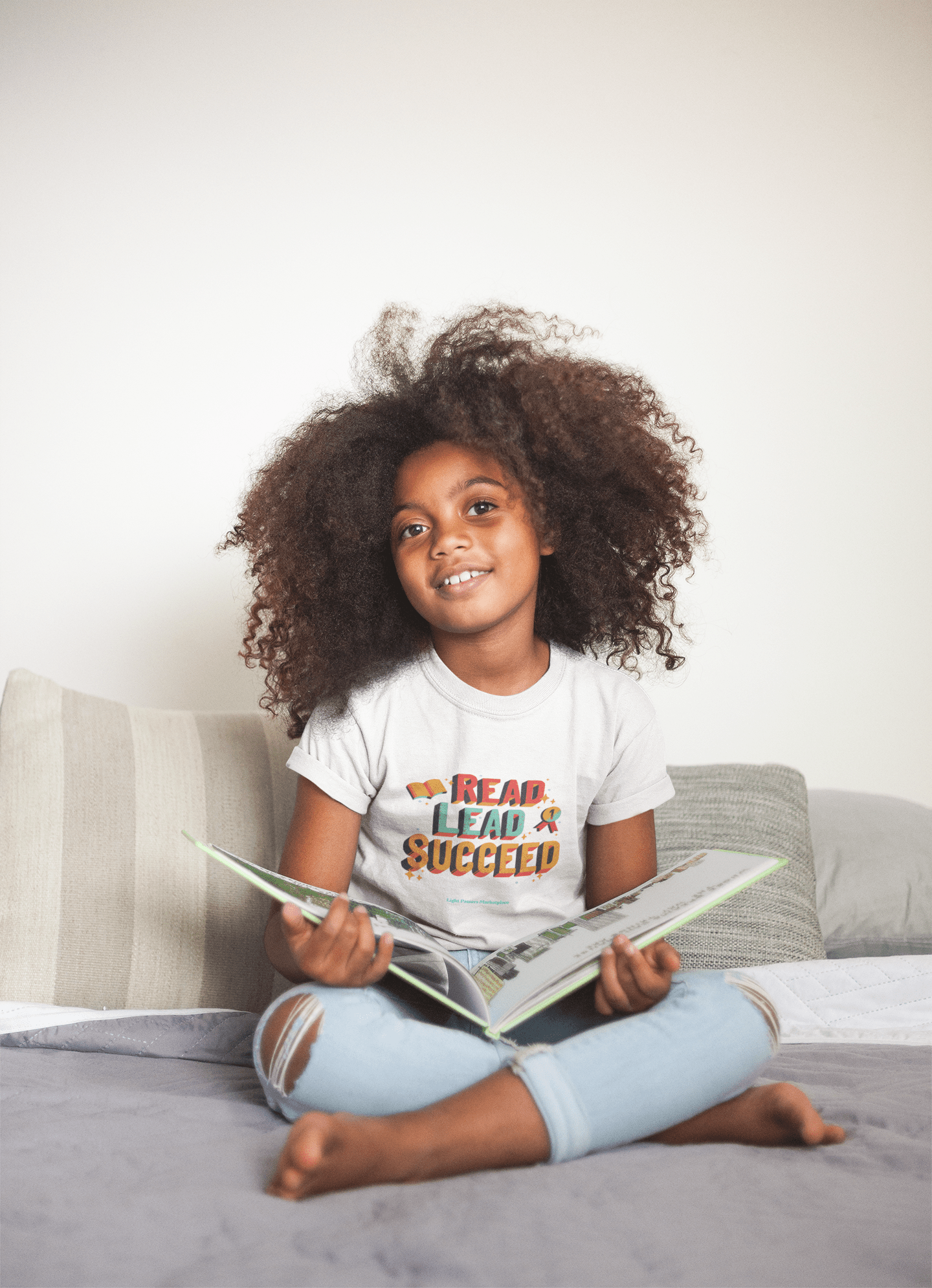 A girl with curly hair reading a book on a couch, showcasing the Read Lead and Succeed Youth T-shirt. Quality 100% cotton tee for kids, featuring durable twill tape shoulders and curl-resistant collar.