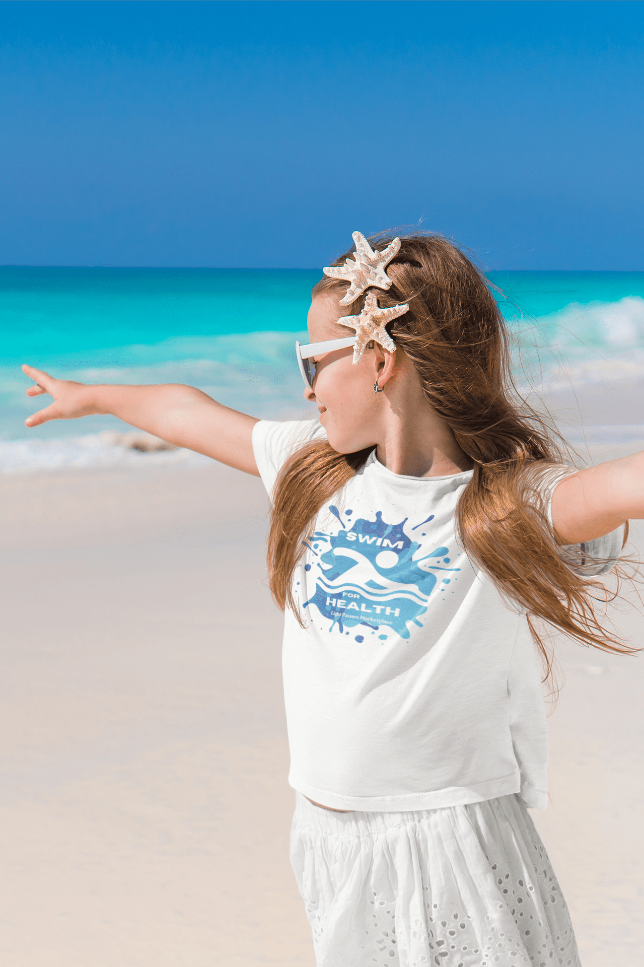 A girl on the beach with arms out, wearing a white shirt. Close-up of a starfish in hair. Product: Swim for Health Youth T-shirt, 100% cotton, ribbed collar, tear-away label, ethically made.