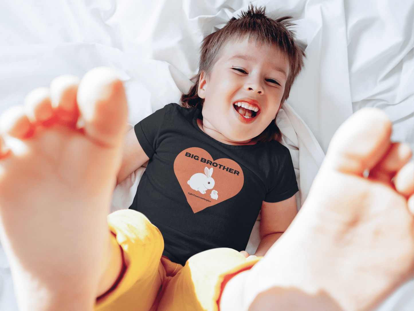 A smiling boy in a Big Brother Toddler T-shirt, lying down with feet up. Soft, 100% combed cotton tee, gentle on sensitive skin, featuring a heart and rabbit cartoon print.