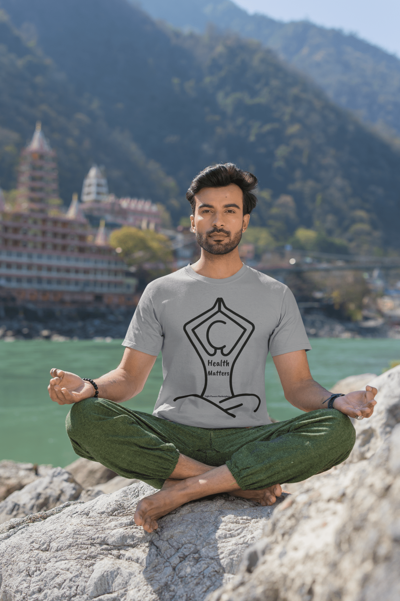 A man in a soft-style unisex t-shirt sits on a rock by a body of water. Made of 100% cotton, this tee offers comfort and durability with twill tape shoulders and ribbed collar.