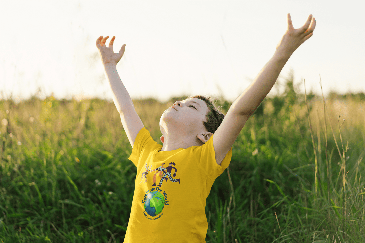 A boy in a yellow shirt with arms raised, showcasing the Celebrate Our World Youth T-shirt. Made of 100% cotton, featuring durable twill tape shoulders and curl-resistant collar. Ethically sourced US cotton.