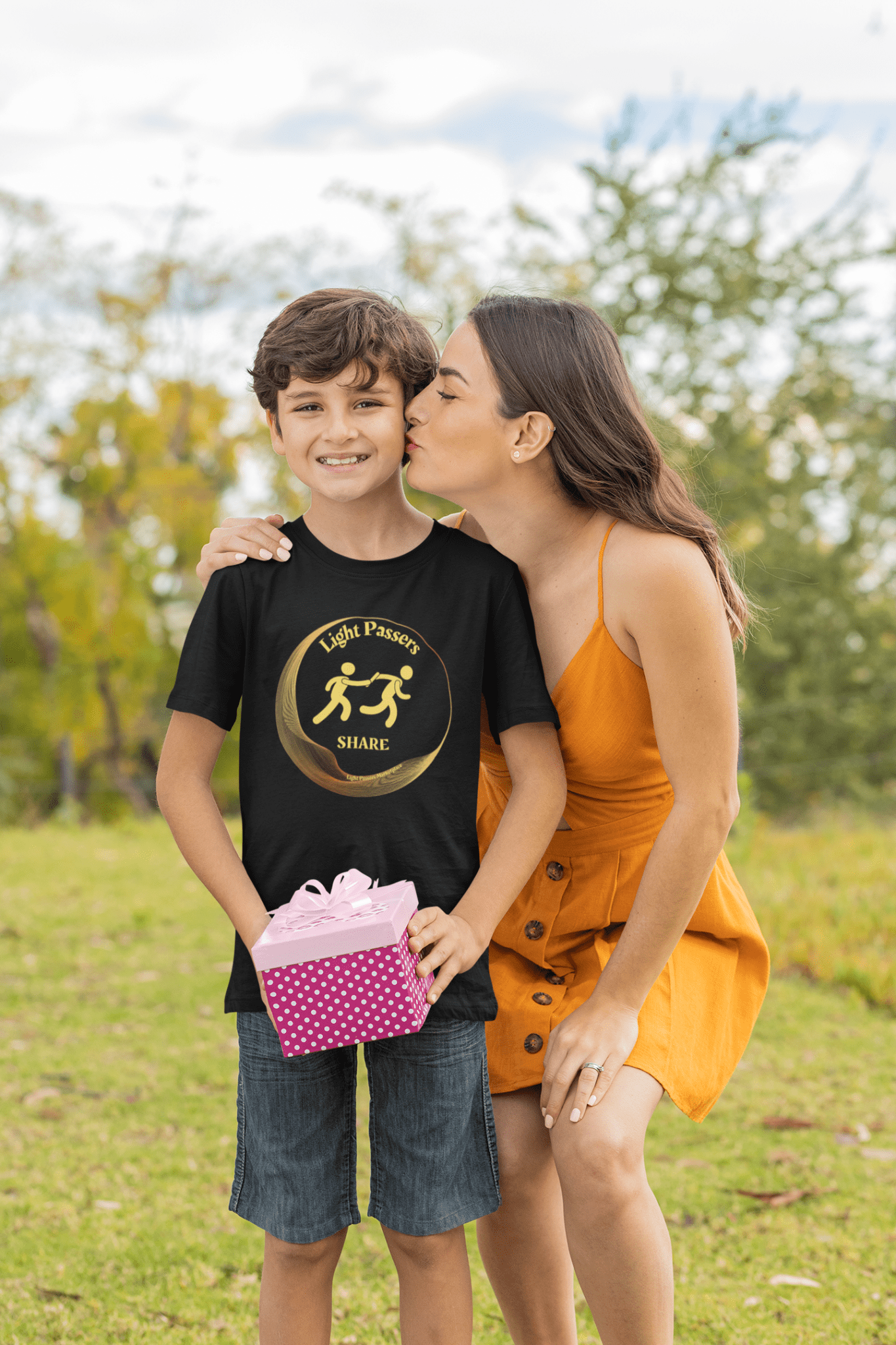Woman kisses boy's cheek as he wears The Light Love and Peace Light Passers Share Youth T-Shirt, holding a pink and white gift box.