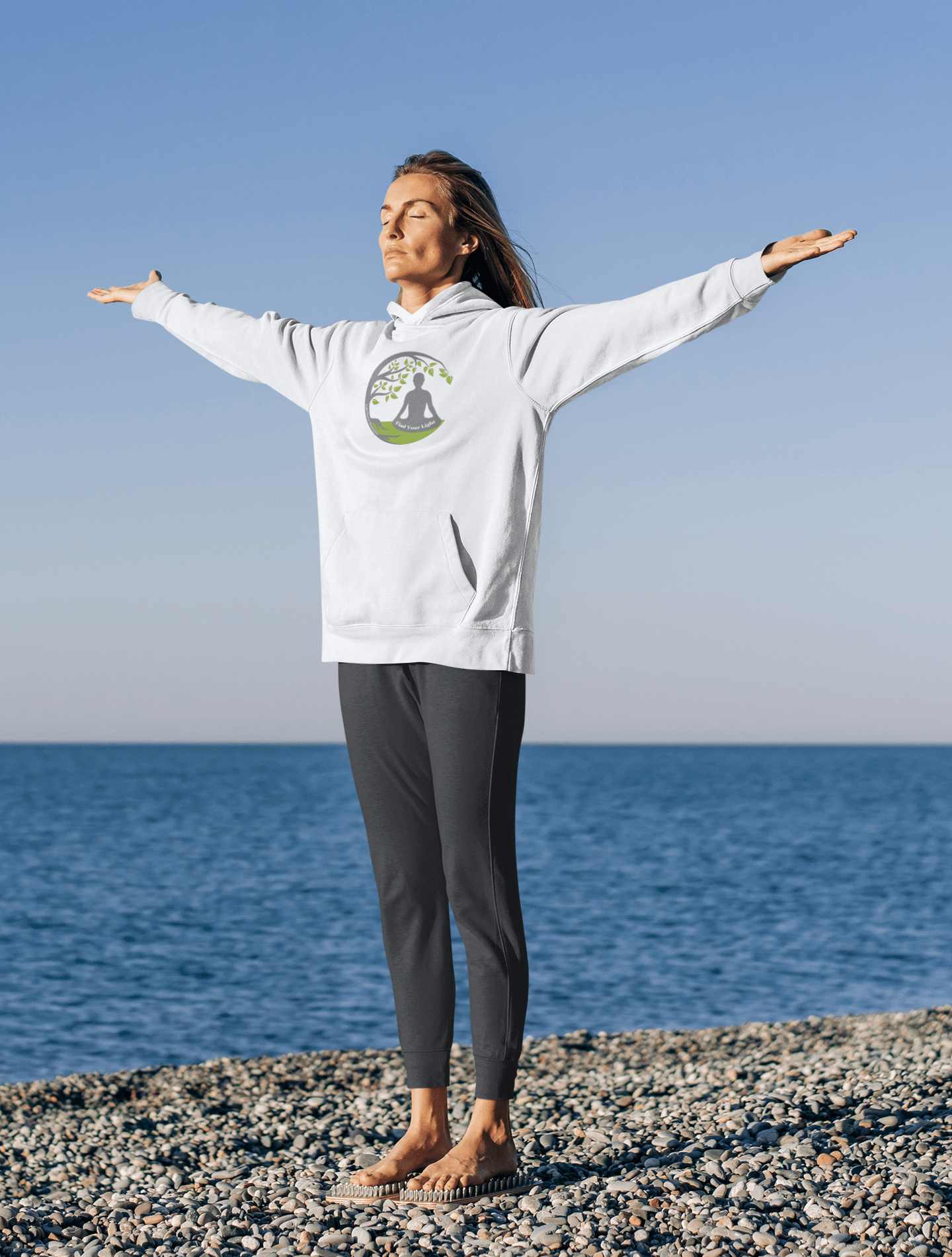 Woman on beach wearing Health The Light Yoga Meditation Adult Unisex Hoodie, arms outstretched, showcasing comfort and practical design with kangaroo pocket and color-matched drawstring.