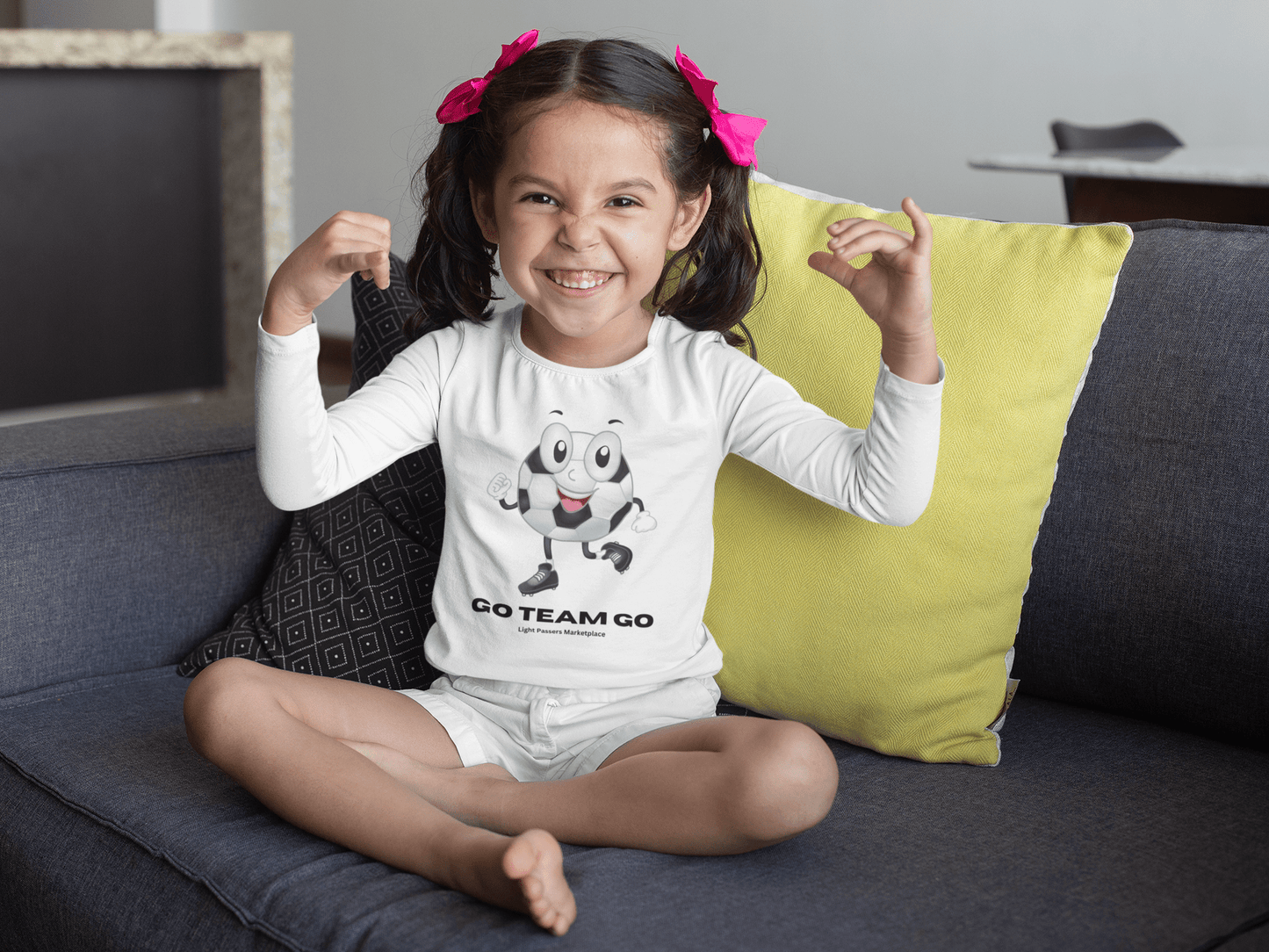 Toddler wearing Soccer Ball Guy Long Sleeve with cartoon soccer ball, sitting on a couch, smiling. Ideal for young soccer fans and game day fun.