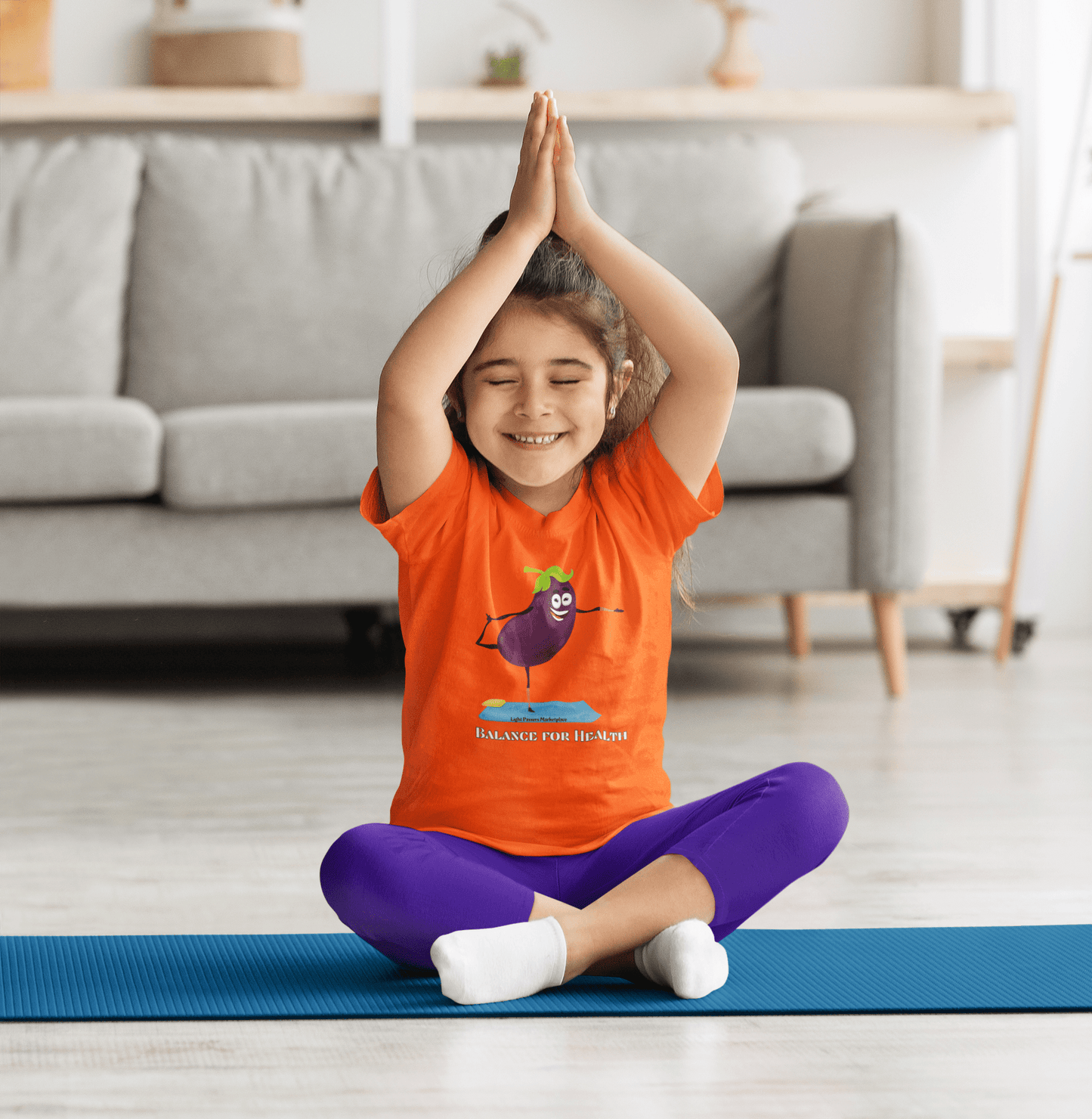 A girl in an orange shirt sits on a yoga mat with hands over head, embodying balance and health. Eggplant Balance for Health Toddler T-shirt, soft 100% cotton, durable print, tear-away label, true to size.