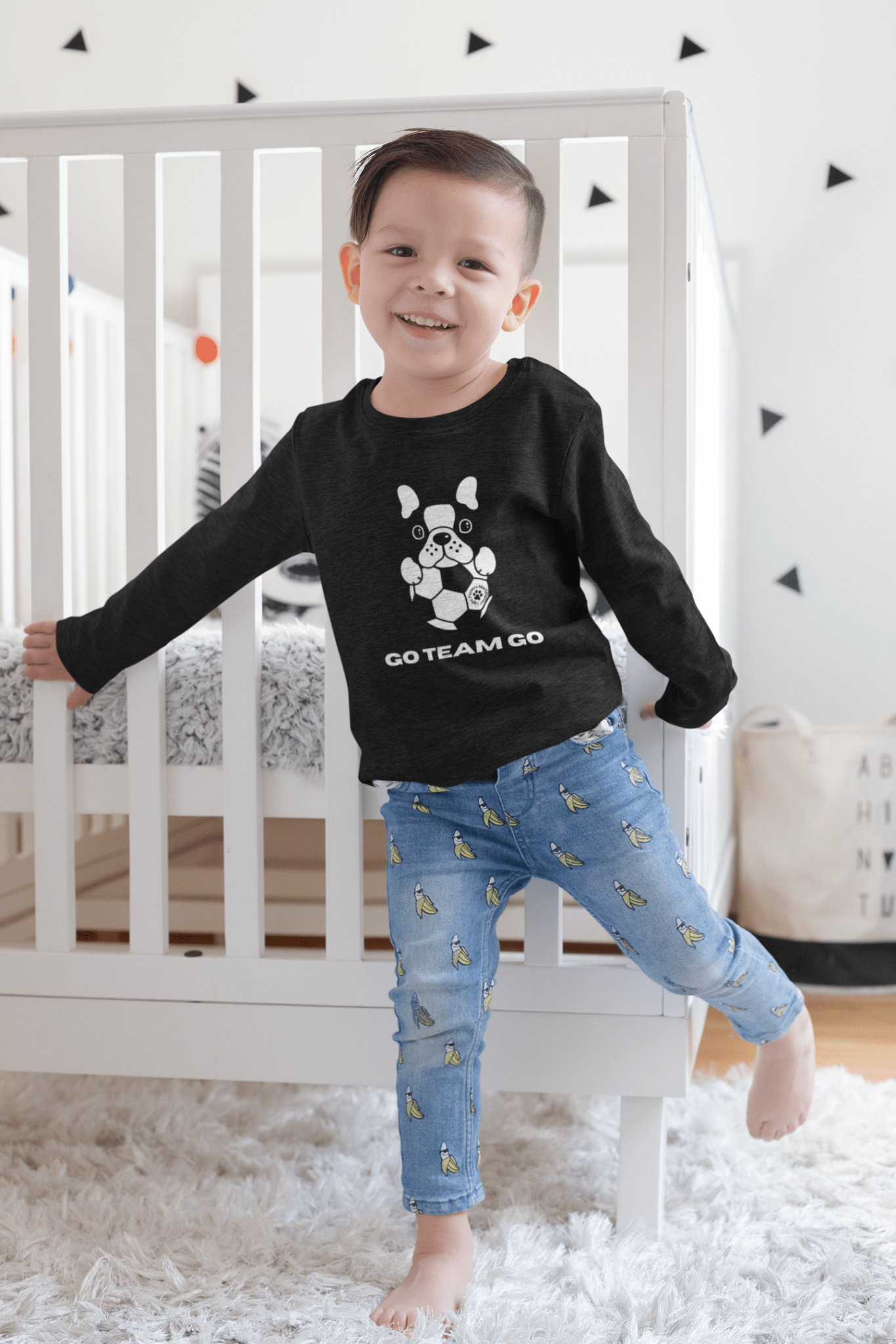 Toddler wearing Soccer Dog Toddler Long Sleeve Tee, featuring a dog design, standing in front of a crib, showcasing its comfortable fit and playful theme.