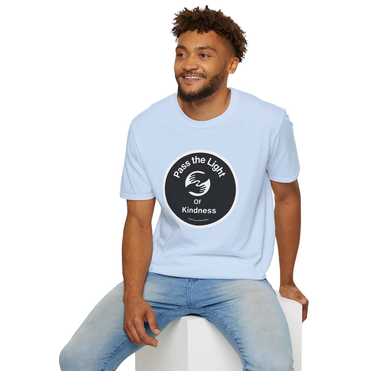 A man in jeans sits on a cube, wearing a Pass the Light of Kindness 2 hands Unisex T-shirt. Close-up of the tee's smooth fabric, ideal for vibrant prints, with no side seams for comfort.