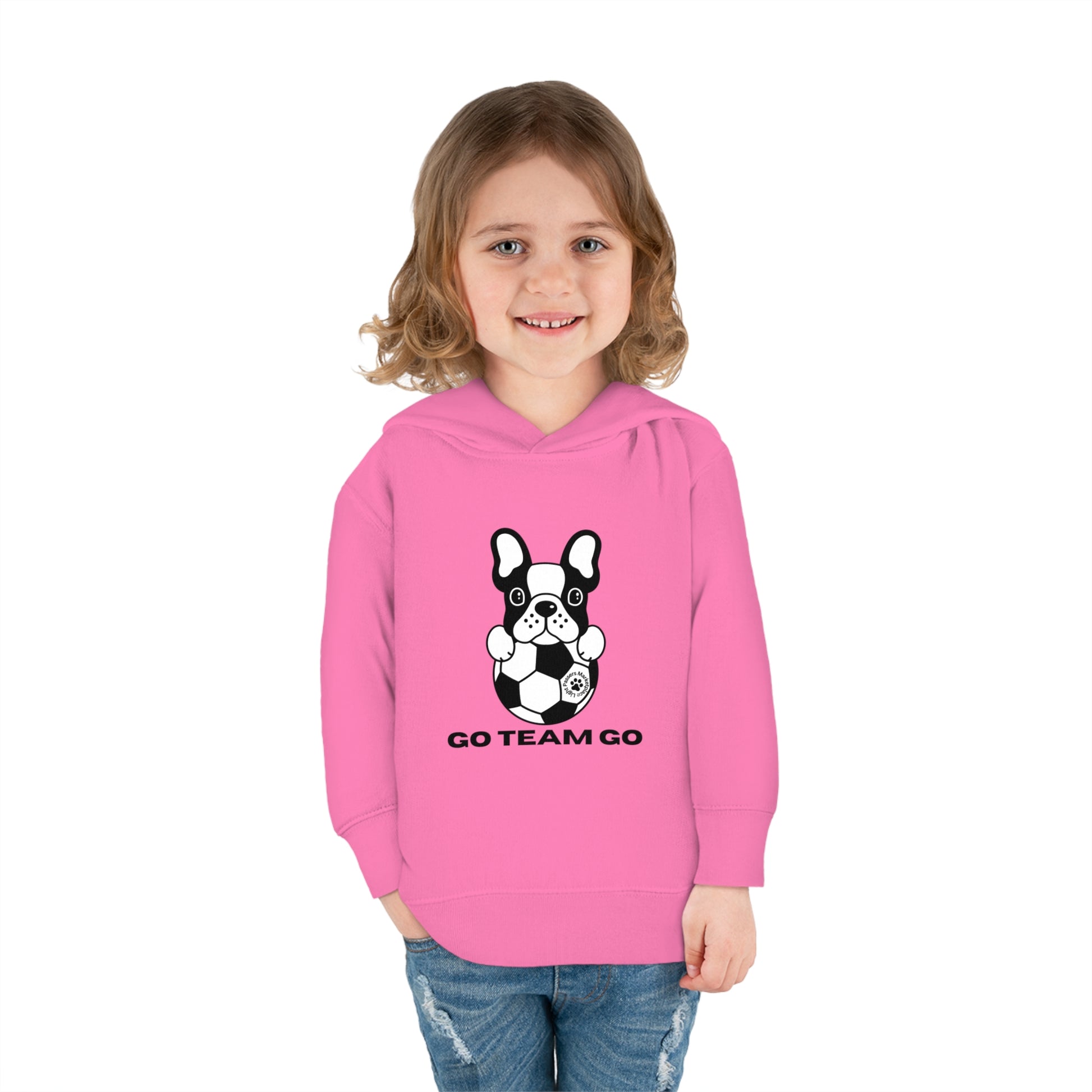 A toddler wearing a Soccer Dog Go Team Go hoodie with a dog and football design. Features jersey-lined hood, cover-stitched details, side seam pockets, and durable cotton-polyester blend for cozy wear.