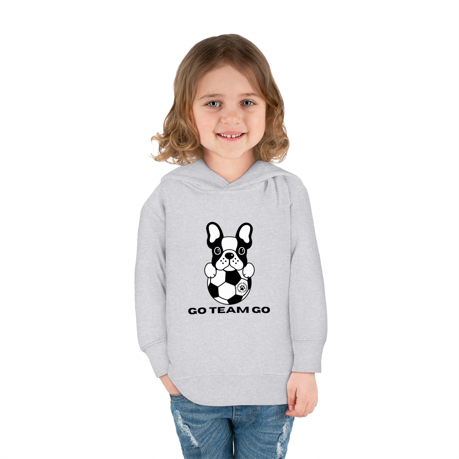 A blonde girl in a grey Soccer Dog Go Team Go Toddler Hooded Sweatshirt smiles, featuring a dog design. Jersey-lined hood, cover-stitched details, and side pockets for comfort and durability.