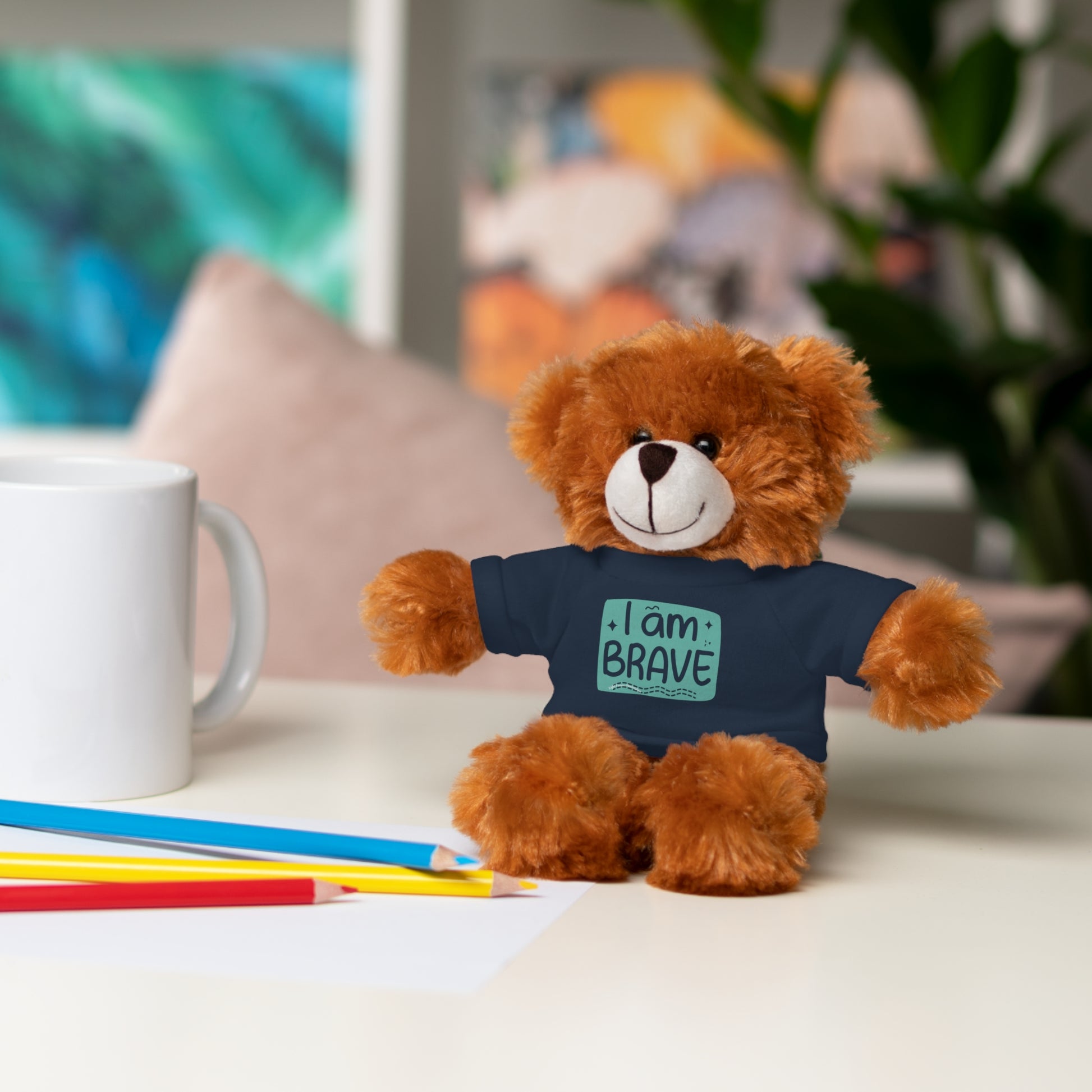 Cool Stuff Stuffies: a teddy bear wearing a removable tee with I am Brave message, posed on a table, next to a white mug.