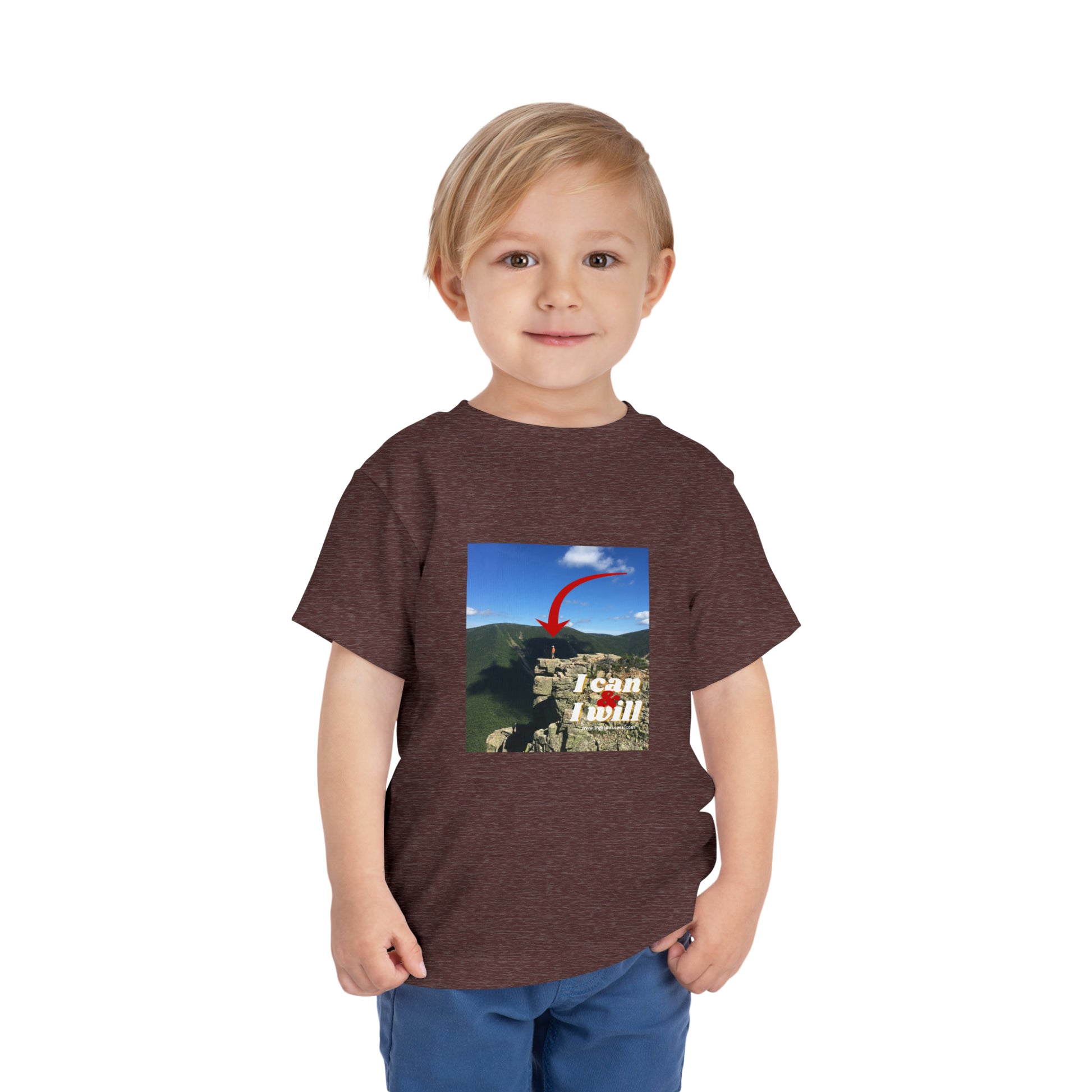 A boy in a brown shirt stands confidently on a cliff, embodying the spirit of adventure on the I Can and I Will Hiking Toddler T-shirt. Made of 100% Airlume cotton for comfort.