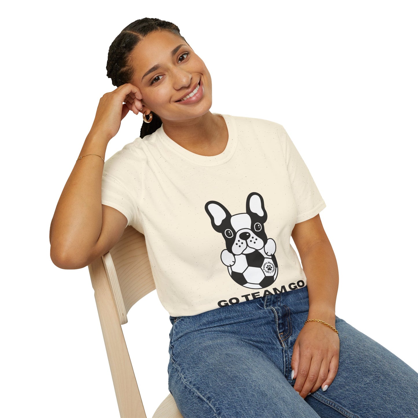 Young person in a Soccer Dog Unisex T-Shirt, featuring a cartoon French bulldog with a soccer ball, smiling while seated on a wooden chair.