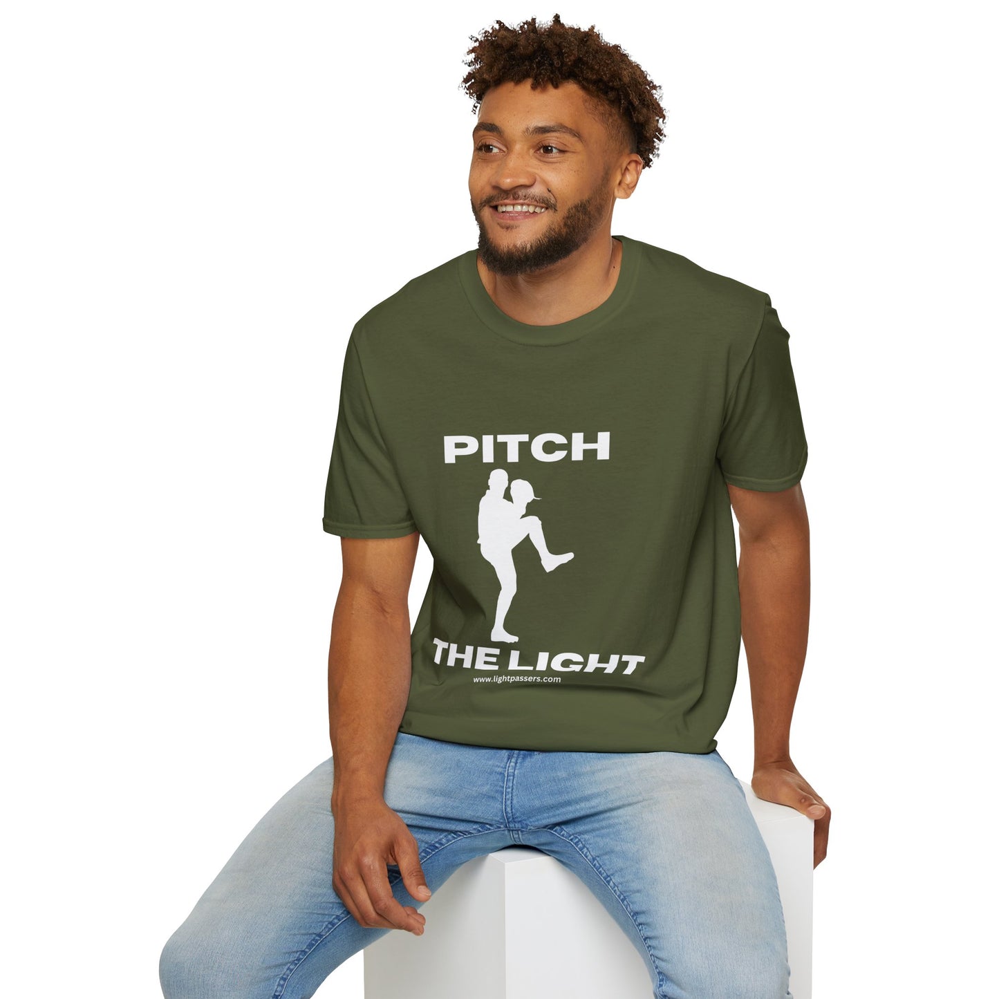 A man in a green shirt sits on a cube, showcasing the PITCH The LIGHT white lettering Unisex T-Shirt. Soft 100% cotton tee with twill tape shoulders, no side seams, and ribbed collar.