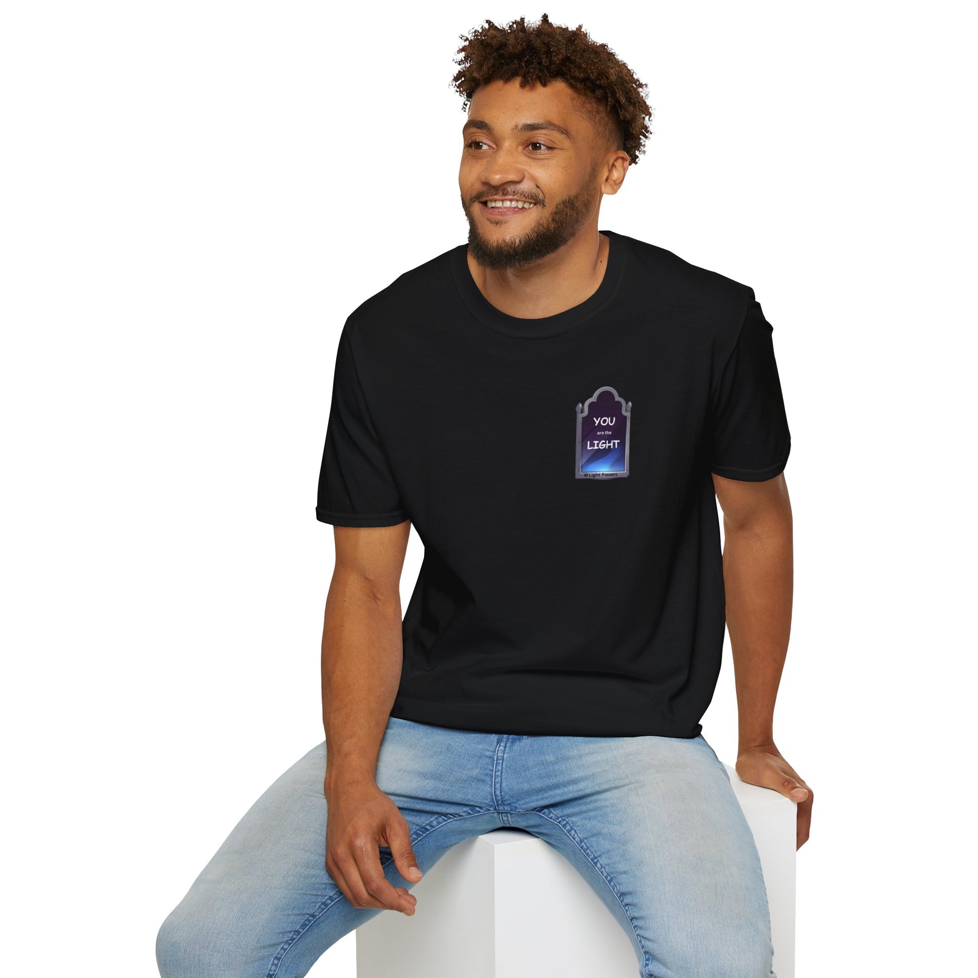 A man in a black shirt sits on a cube, showcasing the YOU ARE THE LIGHT small Mirror Unisex T-shirt. The tee offers a classic fit, 100% cotton fabric, and tear-away label for comfort and style.