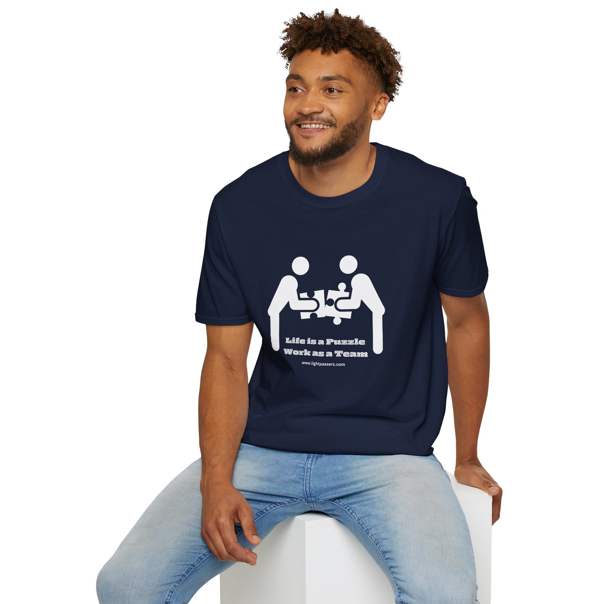 A man in a blue shirt sits on a white cube, showcasing the Life is a Puzzle Team Unisex T-shirt. The tee offers a classic fit, 100% cotton fabric, and tear-away label for comfort and style.