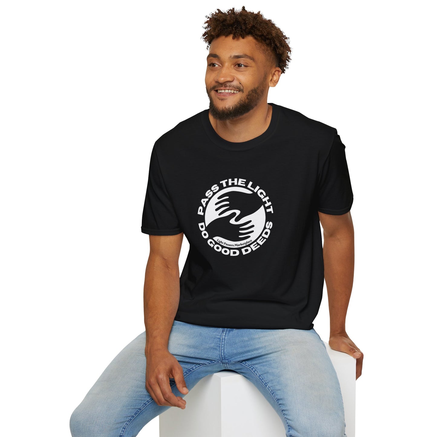 A man in a black shirt sits on a cube, showcasing the Pass the Light Do Good Deeds Unisex T-shirt. The tee features 100% cotton, no side seams, and tape on shoulders for durability.