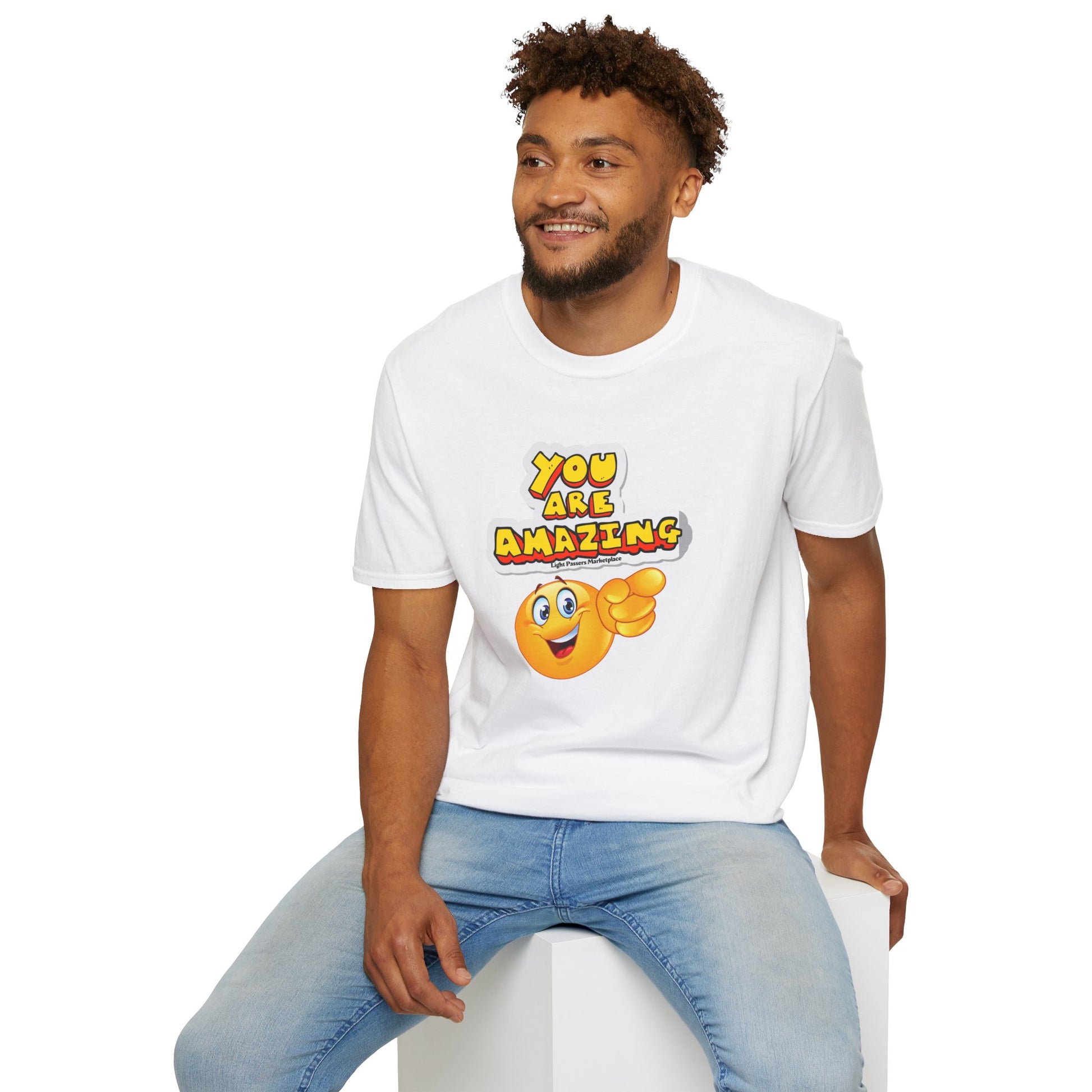 A man in a soft-style unisex t-shirt, sitting on a cube, with a yellow cartoon face pointing. Made of 100% ring-spun cotton, twill tape shoulders, and tear-away label for comfort.