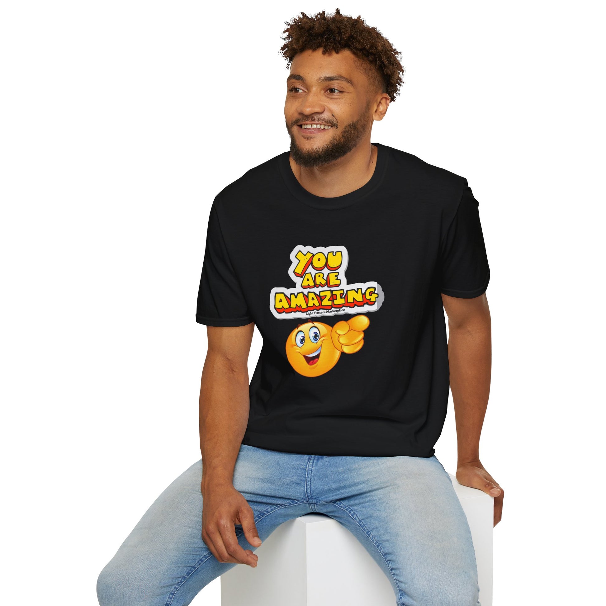 A man in a black shirt sits on a white cube, smiling. Unisex soft-style tee, 100% cotton, lightweight, with twill tape shoulders for durability and a clean crew neckline.