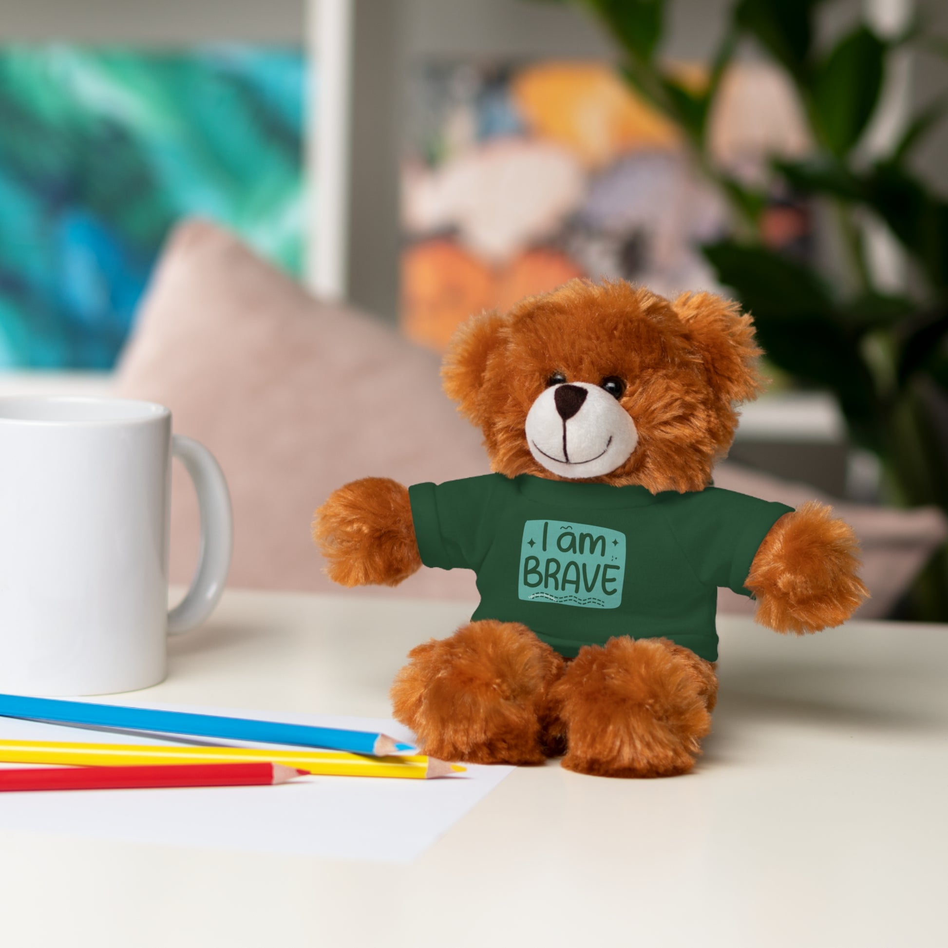Cool Stuff Stuffies plush toy, a teddy bear wearing a customizable green shirt with I am Brave message, next to a white mug on a table.