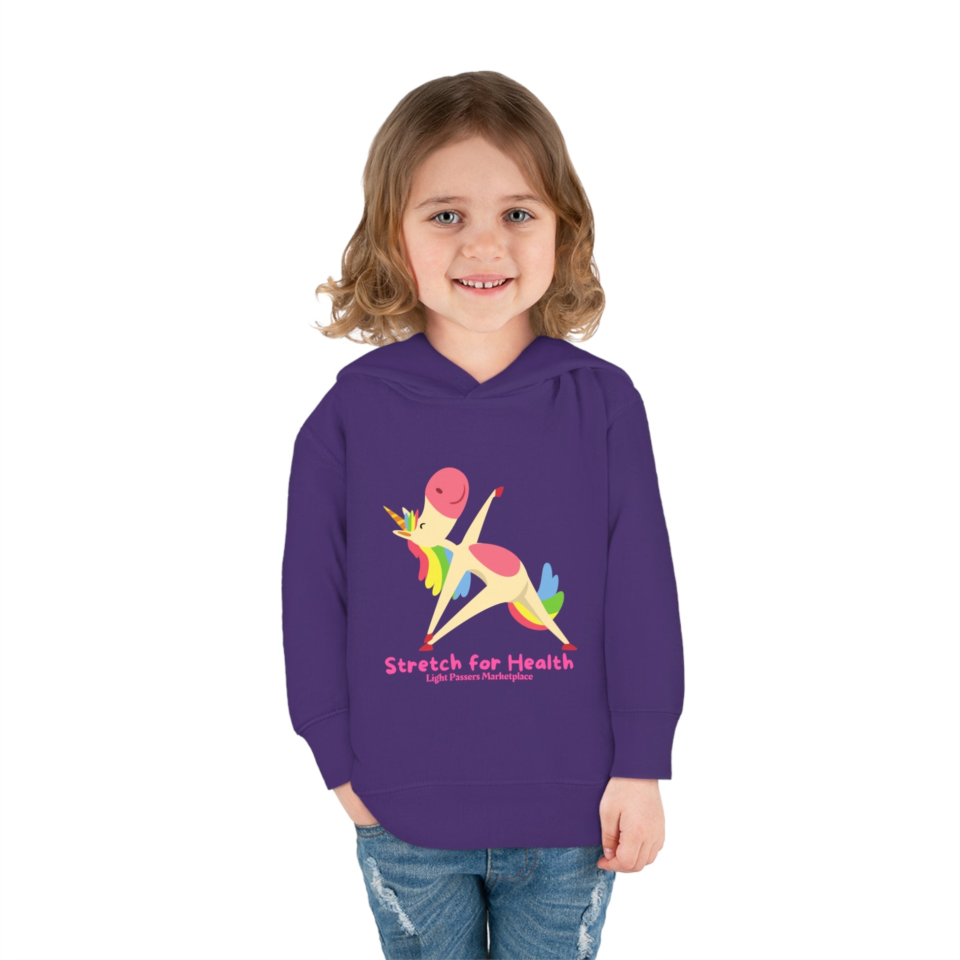 A smiling girl in a purple Rabbit Skins toddler hoodie with a unicorn design, showcasing jersey-lined hood, cover-stitched details, and side seam pockets for durable coziness.
