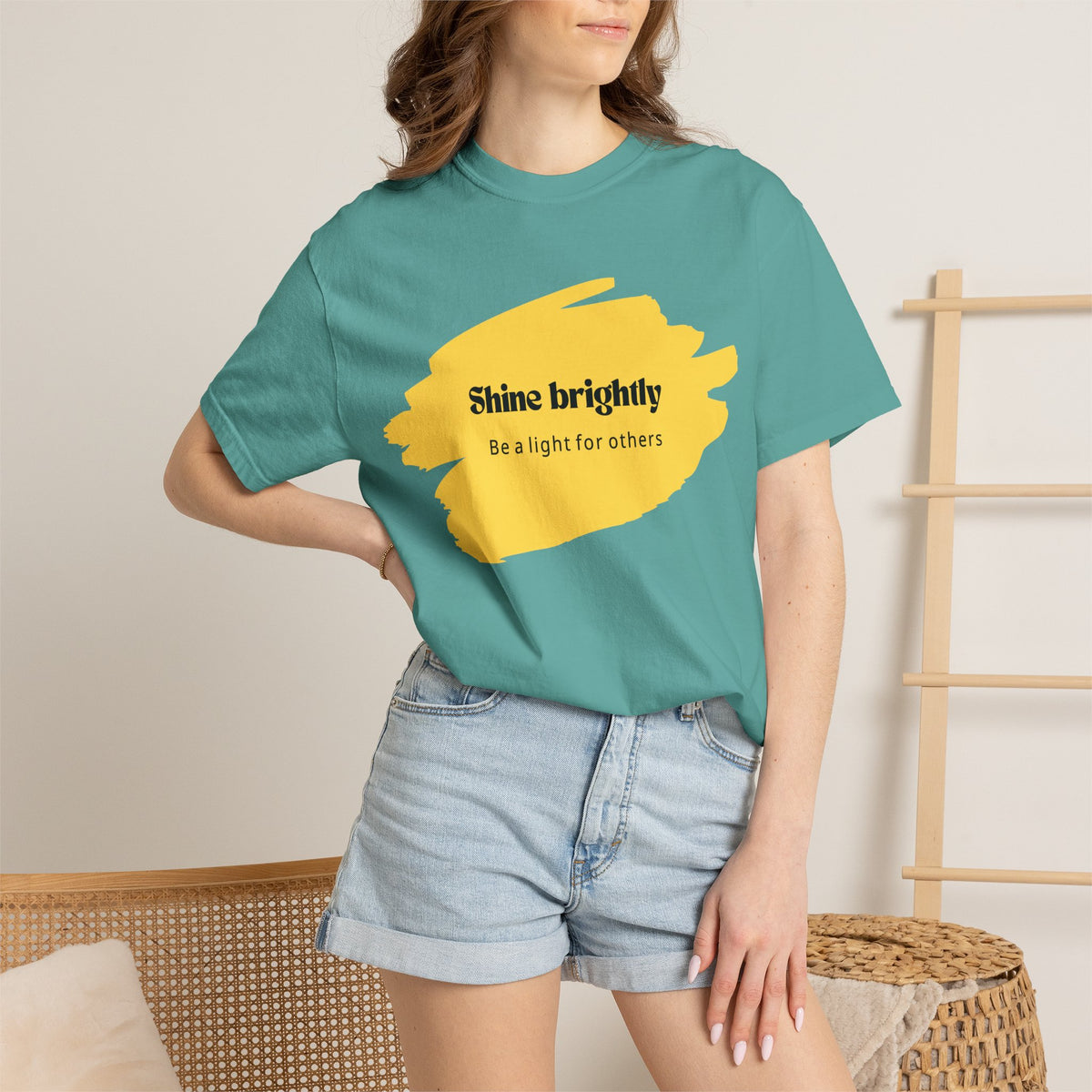 Teal unisex T-shirt worn by model with yellow “Shine brightly” graphic center.