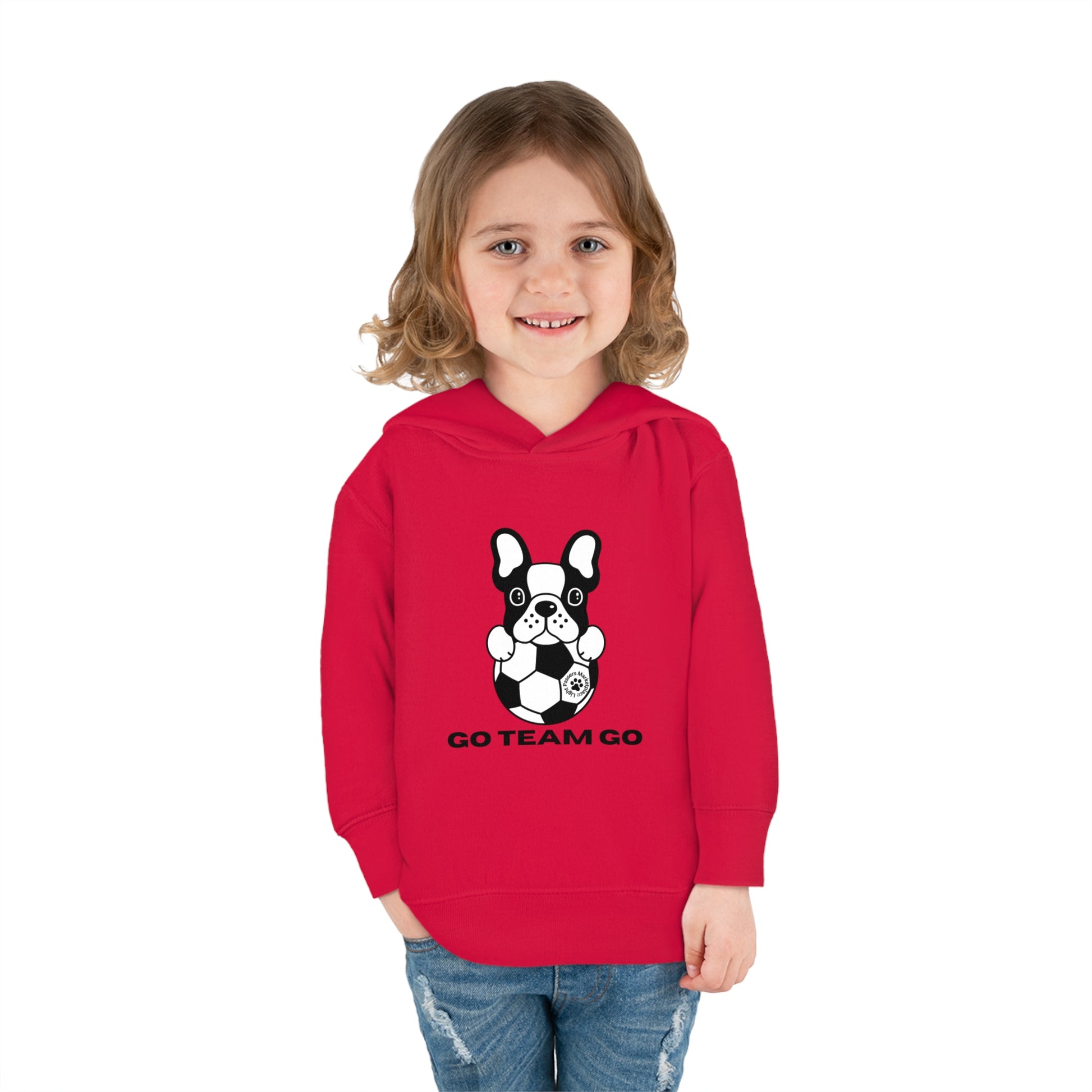 A toddler wearing a red Soccer Dog Go Team Go hooded sweatshirt, featuring a dog design. Made of 60% combed cotton and 40% polyester, with a jersey-lined hood and side seam pockets for comfort.