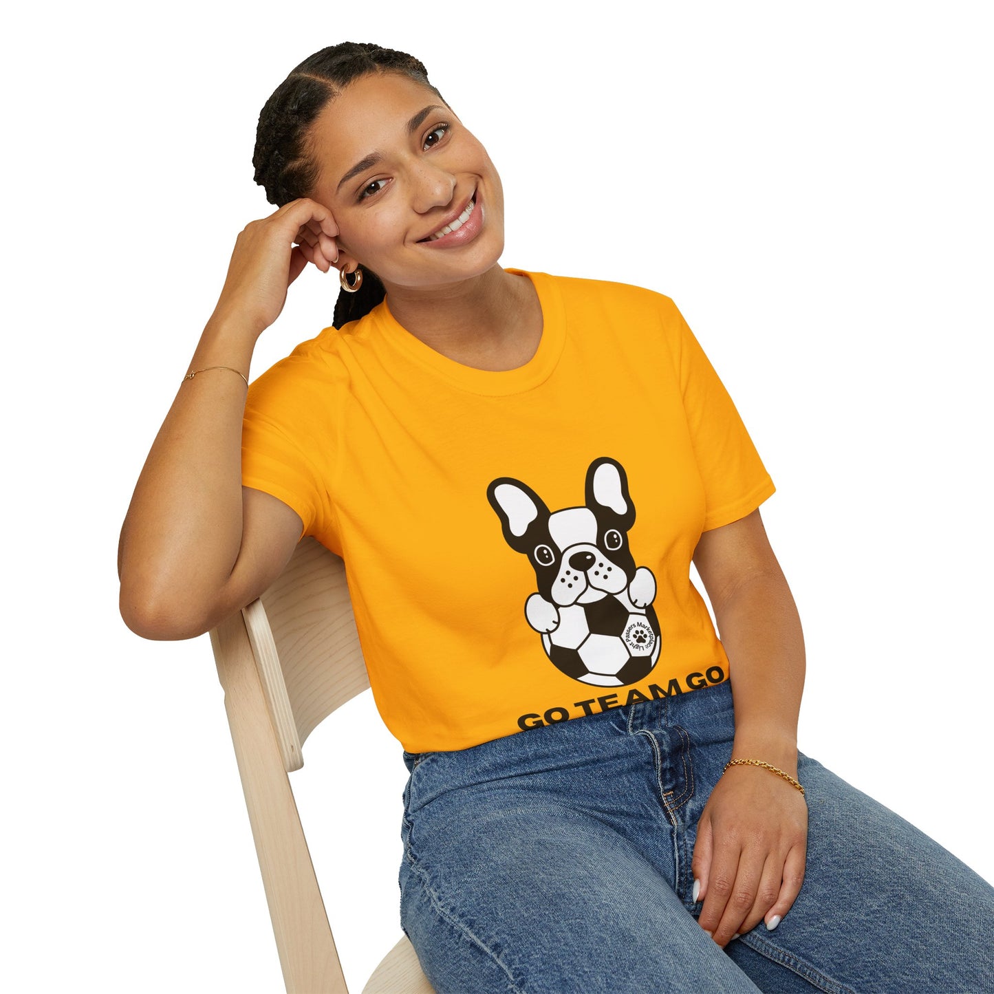 Young woman in a Soccer Dog Unisex T-Shirt with a cartoon French bulldog holding a soccer ball, smiling, wearing jeans and gold jewelry, seated casually.