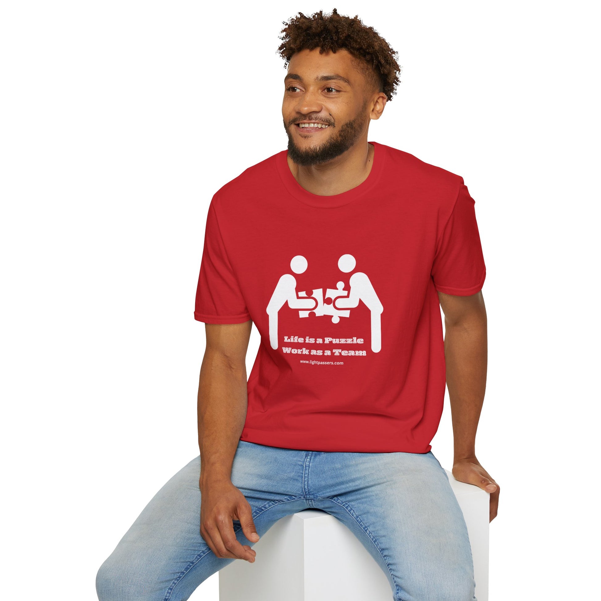 A man in a red shirt sits on a cube, showcasing the Life is a Puzzle Team Unisex T-shirt. The tee features a smooth surface for vivid printing, no side seams, and tape on the shoulders for durability.