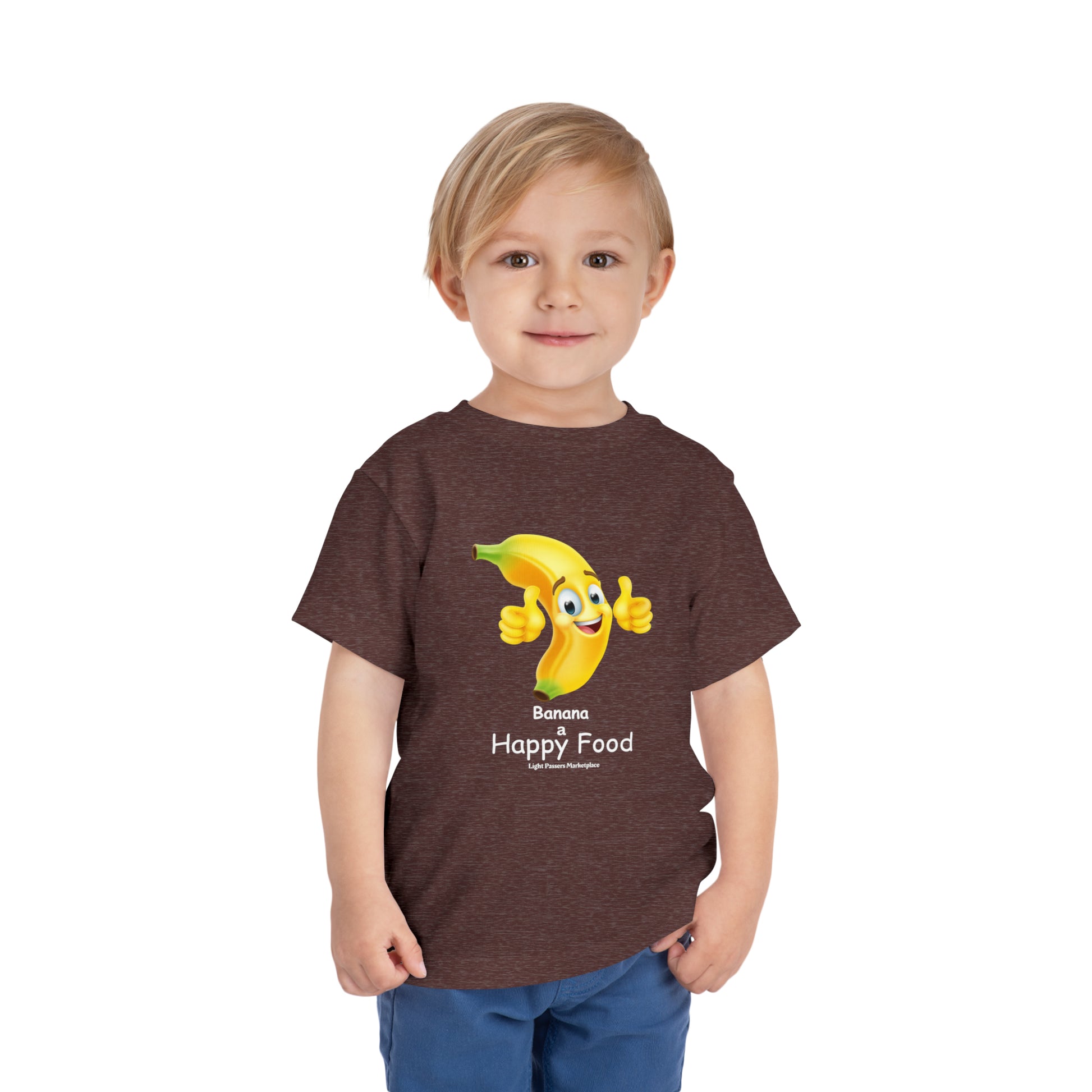 A toddler wearing a brown Banana Happy Food t-shirt, featuring a cartoon banana giving a thumbs up. Made of 100% Airlume combed cotton for comfort.