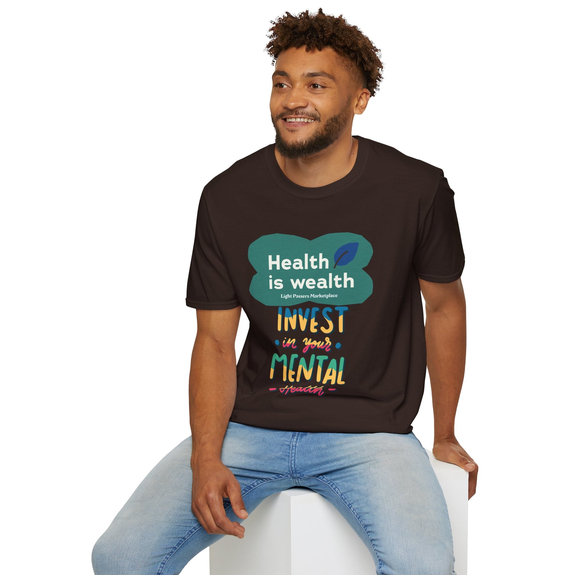 A man in a brown shirt sits on a white cube, wearing a t-shirt with text. Invest in Mental Health Unisex T-shirt: 100% cotton, twill tape shoulders, ribbed collar, tear-away label, ethically made.