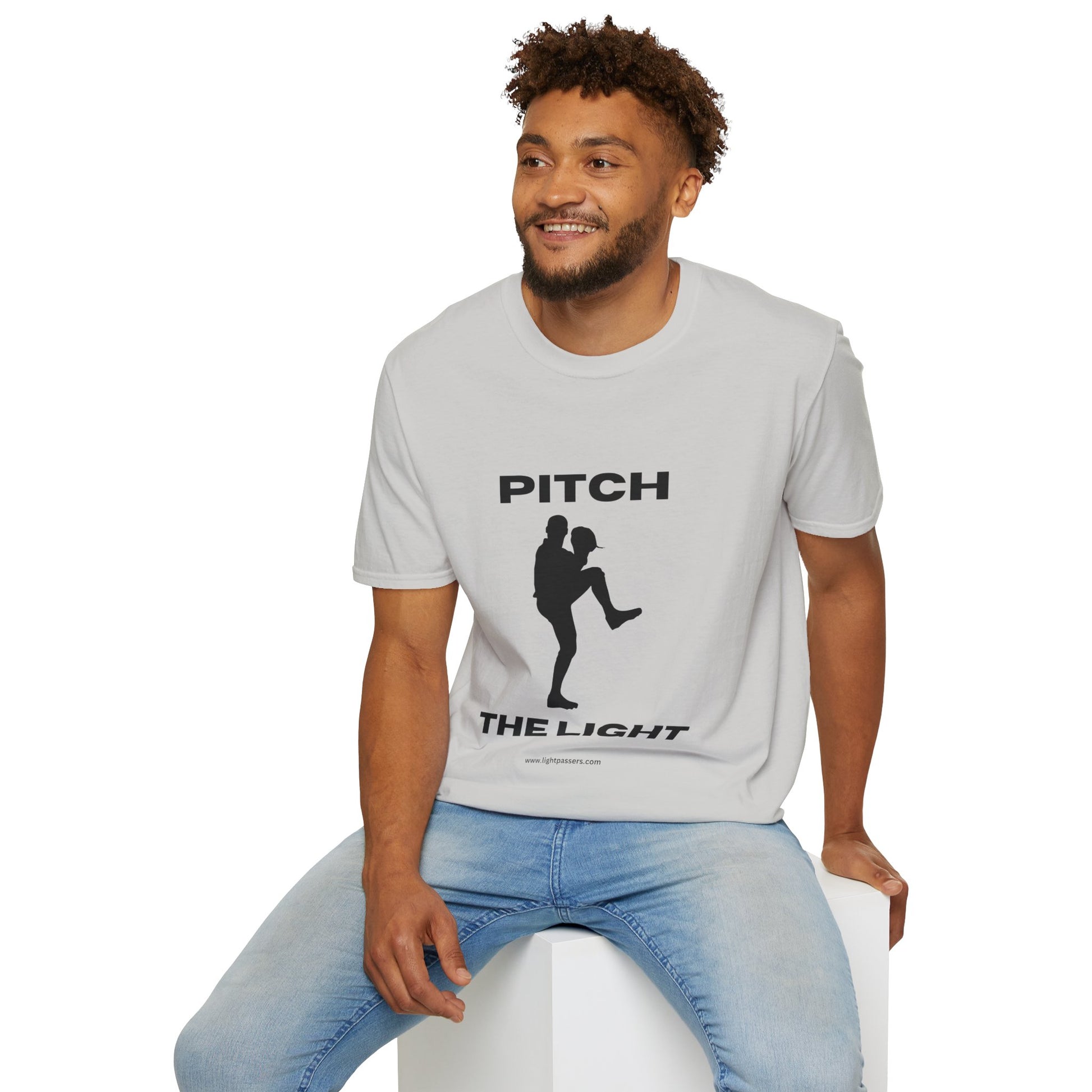 A man sits on a cube, wearing a soft-style PITCH The LIGHT white lettering Unisex T-Shirt. The tee features twill tape shoulders and ribbed collar, made of 100% cotton.