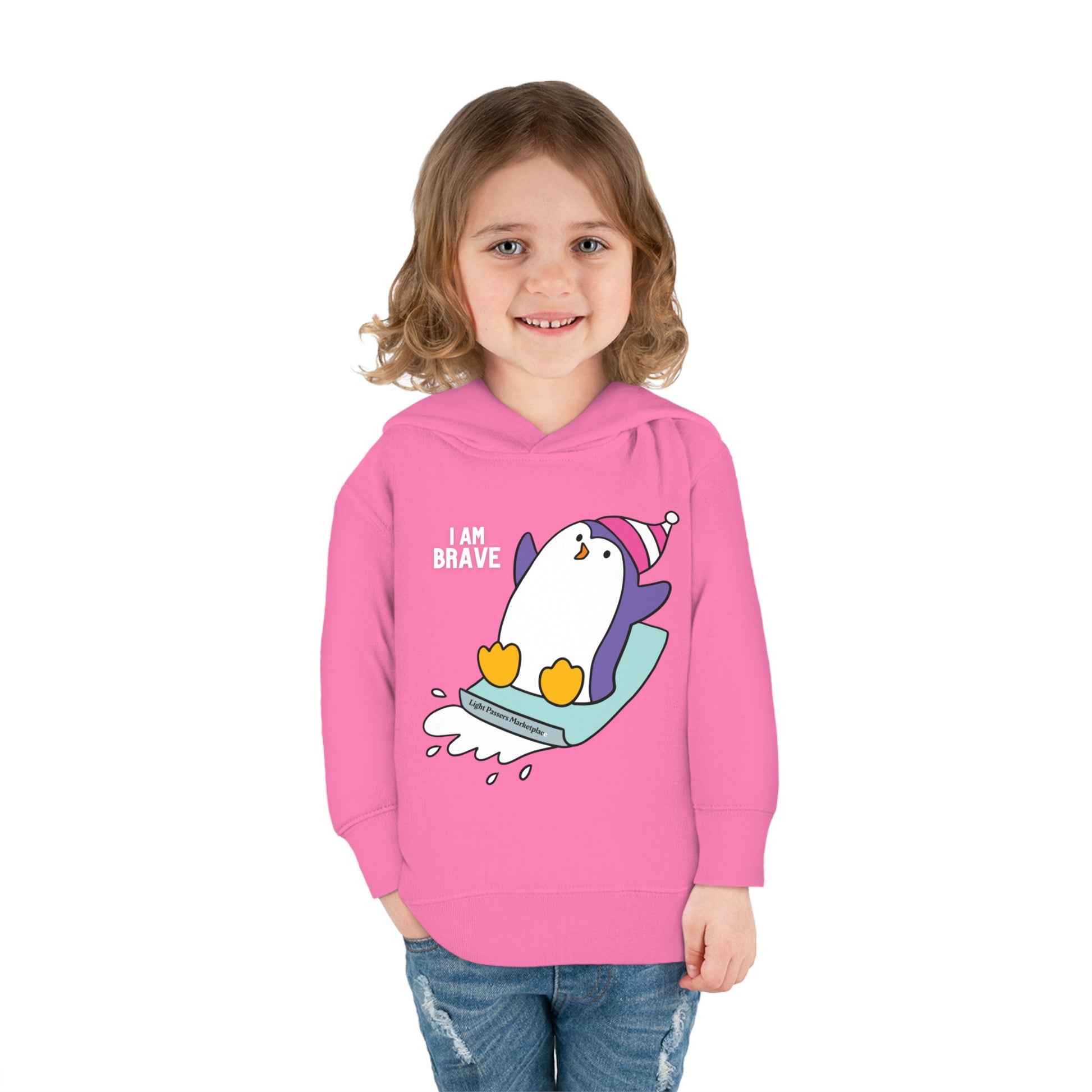 A smiling girl in a Rabbit Skins Brave Penguin Toddler Hooded Sweatshirt with jersey-lined hood, cover-stitched details, and side seam pockets for cozy durability.