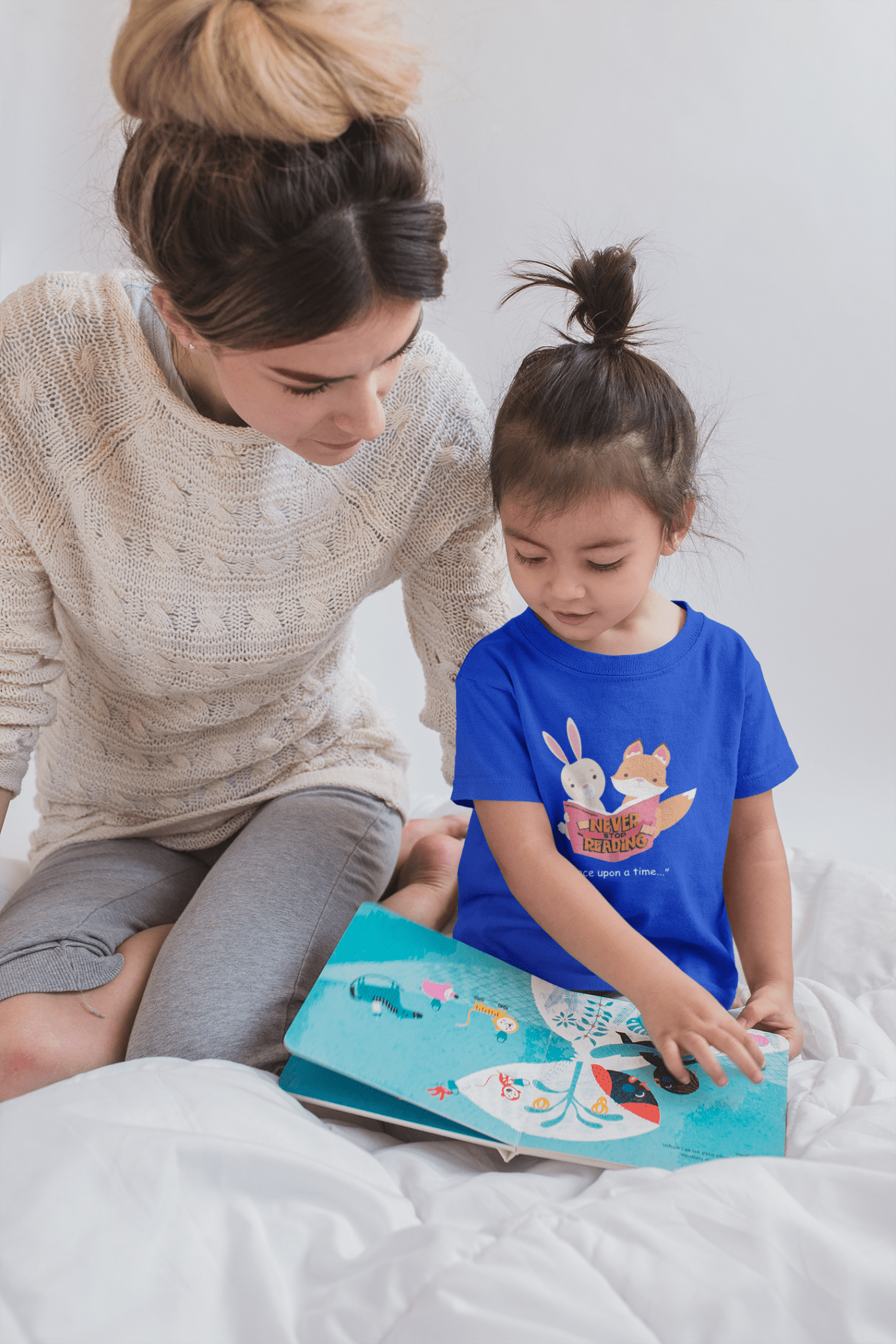 A toddler in a blue shirt reading a book, accompanied by a woman, on a bed. Never Stop Reading Toddler T-shirt: soft, durable, 100% cotton tee for sensitive skin.