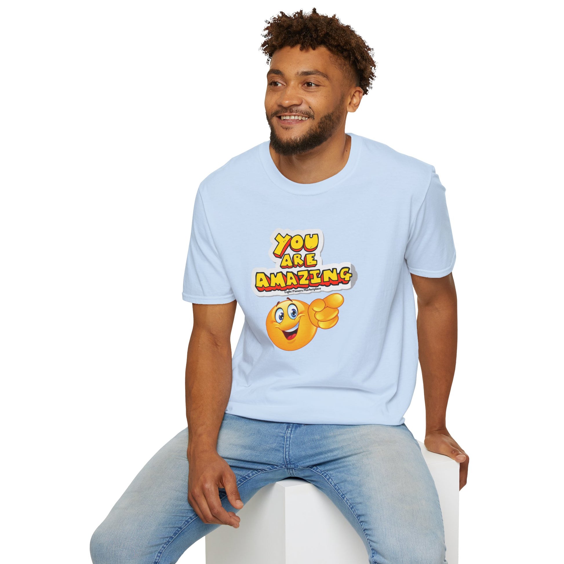 A man in a white shirt sits on a cube, a yellow cartoon character points, and a close-up of a sticker. Unisex You are Amazing emoji pointing T-shirt, 100% cotton, durable twill tape shoulders, ribbed collar.