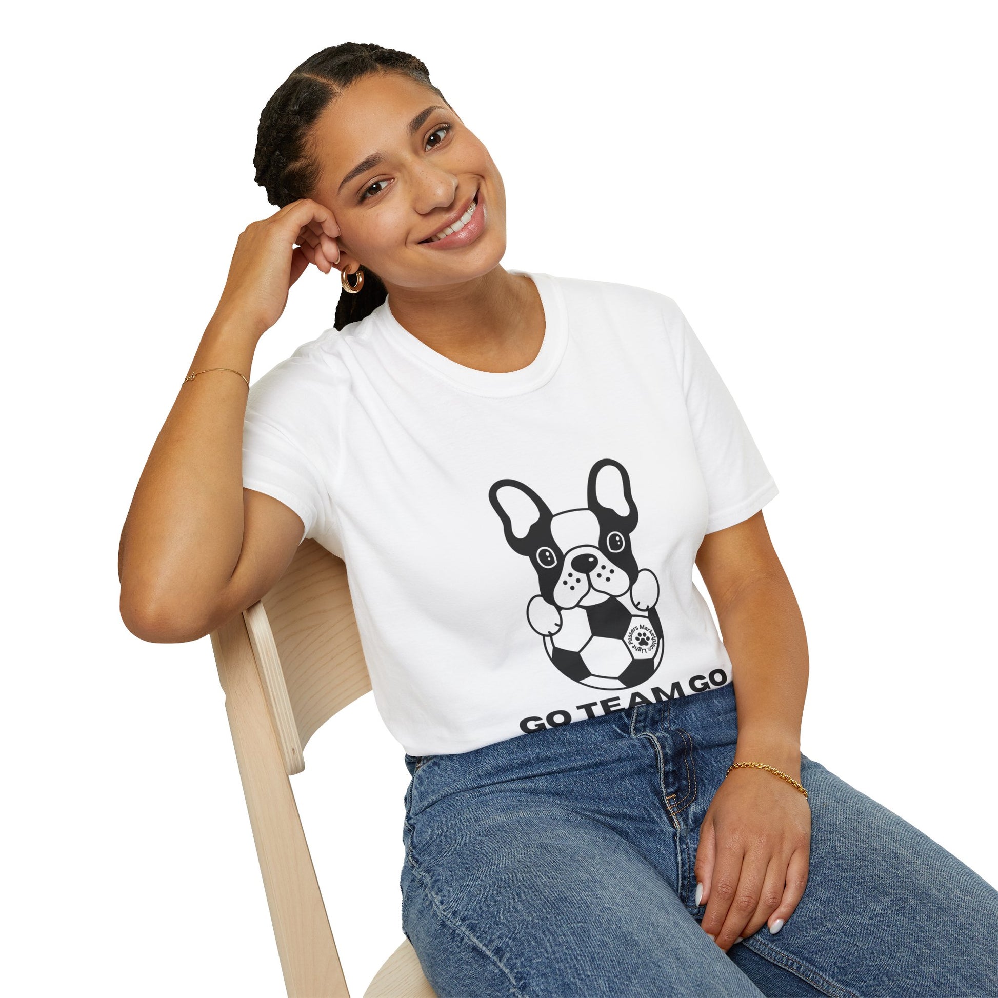 A woman in a white shirt with a dog design sits on a chair, showcasing the Soccer Dog Unisex T-Shirt. The tee, made of soft 100% cotton, offers comfort and durability with twill tape shoulders and a ribbed collar.