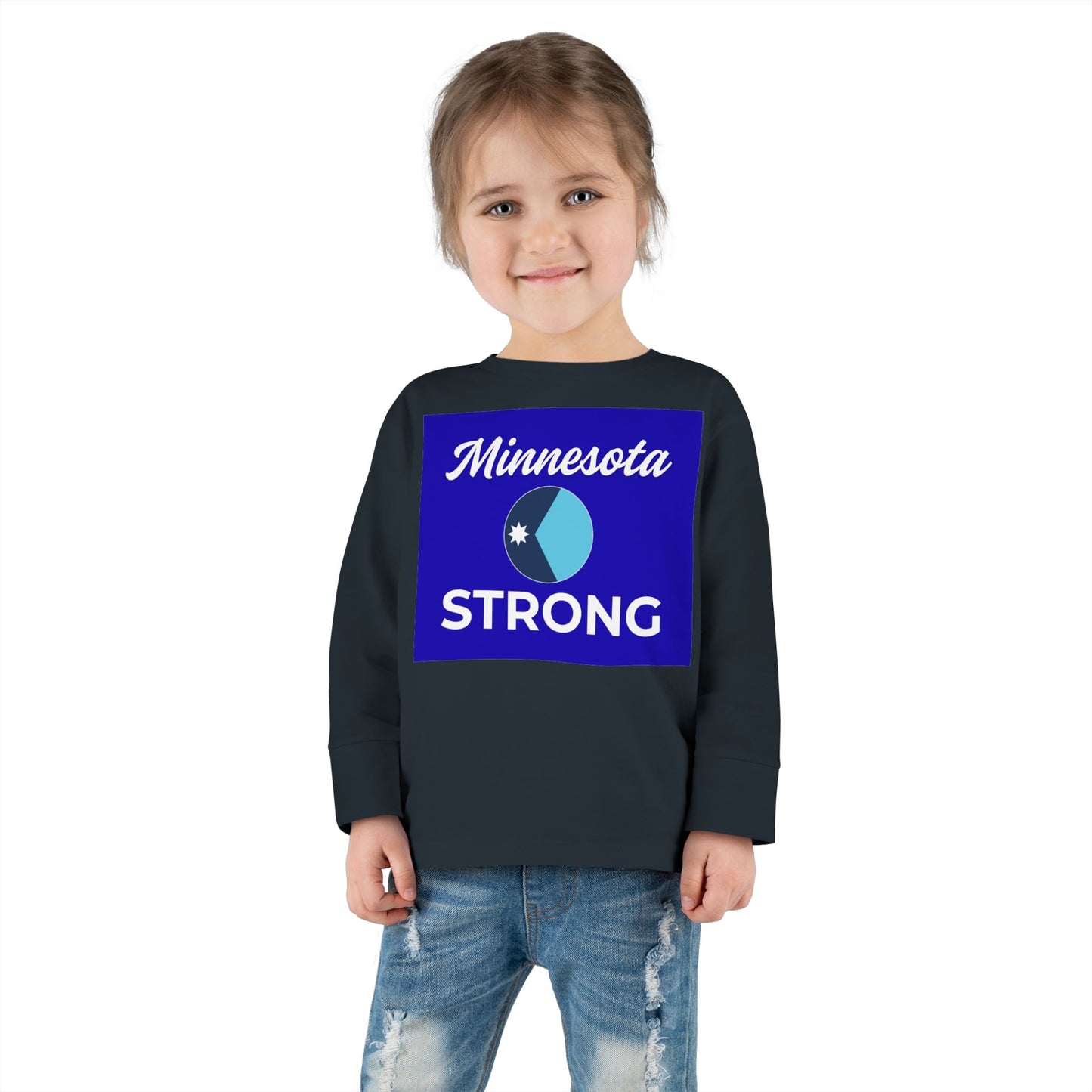 Young girl wearing the Minnesota Strong toddler long sleeve tee, showcasing community pride with a navy shirt and distressed jeans, smiling softly.